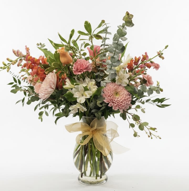 Tatum - A loose, airy bouquet of mixed-color blooms greets the room with garden-fresh charm-perfect as an anniversary or just-because flower arrangement that feels both celebratory and effortless. The pink, white and orange palette reads warm and uplifting, making it an especially nice choice for Spring, a get-well surprise, or a thoughtful housewarming gift that brightens a new space.  This arrangement mixes snapdragons, roses, stock, alstroemeria, carnations, chrysanthemums and lilies with rounded eucalyptus foliage for textural contrast and a slightly wild, hand-gathered silhouette. Tall stems add vertical movement while clustered blossoms provide a full, generous presence; everything sits in a clear glass vase tied with a gold ribbon for a polished, gift-ready finish.  Send this when you want the recipient to feel genuinely seen-whether saying thank you, congratulations, or cheering someone on through recovery. Sized to display on a dining table or bedside, the design arrives ready to enjoy; keep flowers in fresh water, trim stems occasionally, and place away from direct heat to extend their life.