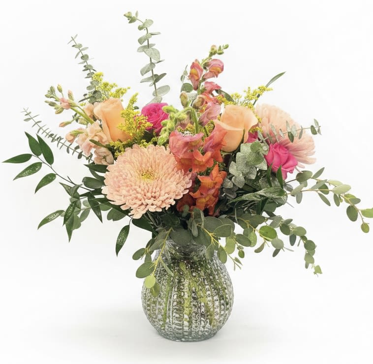 Elodie - Soft peach and bright pink blooms mingle in a loose, garden-style arrangement that feels freshly picked - perfect as a birthday bouquet or a cheerful everyday flower arrangement to lift someone's spirits. The composition reads light and joyful, making it an ideal choice for spring celebrations or to say "I'm thinking of you" in a way that feels both tender and energetic.  A large pale peach chrysanthemum anchors the front while pink roses and tall snapdragon-like spikes create height and movement; alstroemeria and airy yellow filler weave through abundant eucalyptus for soft texture and a subtle herbal scent. The flowers arrive arranged in a rounded green glass vase with a mottled, textured finish that gives the piece a handcrafted, decorative touch and complements the bouquet's pastel palette.  Give this to a friend, mother, or colleague who delights in soft color and natural, garden-inspired style - she'll feel genuinely celebrated and seen. Vase-arranged for immediate display; refresh water as needed and trim stems every few days to extend bloom life.