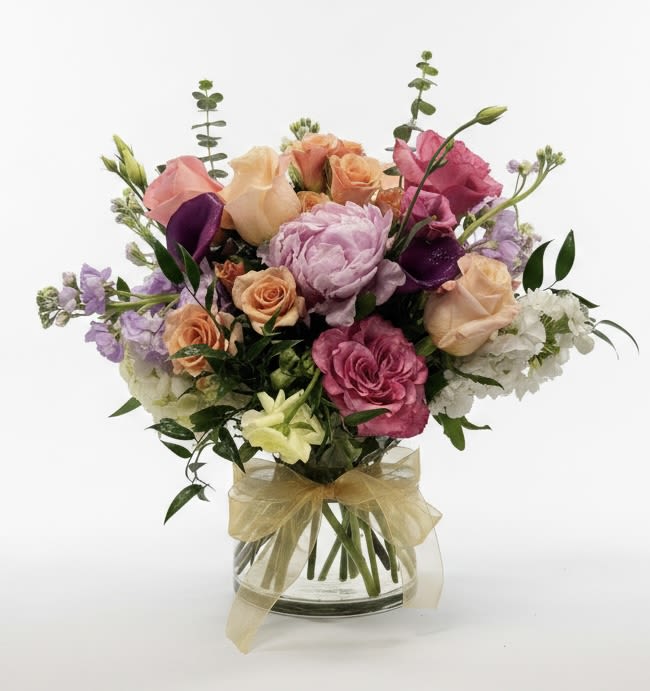 Marlowe - A full, rounded bouquet of pink, peach, purple and white blooms makes an immediate, joyful impression-perfect as a birthday bouquet, anniversary arrangement, or a spring celebration gift. Marlowe pairs romantic roses with soft lisianthus and airy stock to create a garden-fresh look that reads both elegant and effortlessly cheerful; the person you're thinking of will feel noticed and warmly celebrated when these blooms arrive.  The arrangement is loosely gathered in a clear glass vase and finished with a soft gold ribbon at the base for a polished, feminine touch. Tall accent stems rise above the main cluster to add height and a lightly garden-style silhouette, while the mix of textures-from velvety rose petals to the delicate ruffled lisianthus-keeps the composition lively. Color tones move from warm peach to blushing pink with purple and white highlights for a balanced, wearable palette.  Marlowe is an excellent choice for everyday gifting as well as special moments like anniversaries and springtime congratulations; it reads as both intimate and festive. Delivered ready to display in its vase, simply trim the stems as needed and refresh the water every few days to extend the life of the flowers.