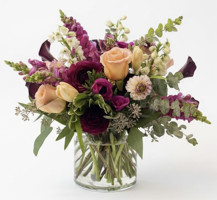 Maeve - A lush, garden-style mixed flower arrangement that feels both effortless and intentional-perfect as a birthday bouquet, a thank-you gift, or a cheerful seasonal display. This flower arrangement for celebrations brings together peach and pink roses, pale tulips, and pops of deep magenta to create an immediate visual warmth that will make the person you're thinking of feel genuinely celebrated.  The composition pairs rounded focal blooms like roses and ranunculus with upright snapdragons and tulip stems for varied height and texture; white daisies and delicate accent flowers brighten the mix while dark plum calla lilies and silvery-green foliage add dramatic contrast and depth. Arranged densely and slightly asymmetrically in a clear glass vase, the piece reads like a freshly gathered garden bouquet with a compact, lush profile.  Ideal for sending to a friend, coworker, or family member who loves vibrant yet refined color, this arrangement suits casual celebrations and everyday moments alike. The clear glass vase is included-no assembly required-and basic care of fresh water changes and keeping stems out of direct heat will help the flowers look their best through the season.