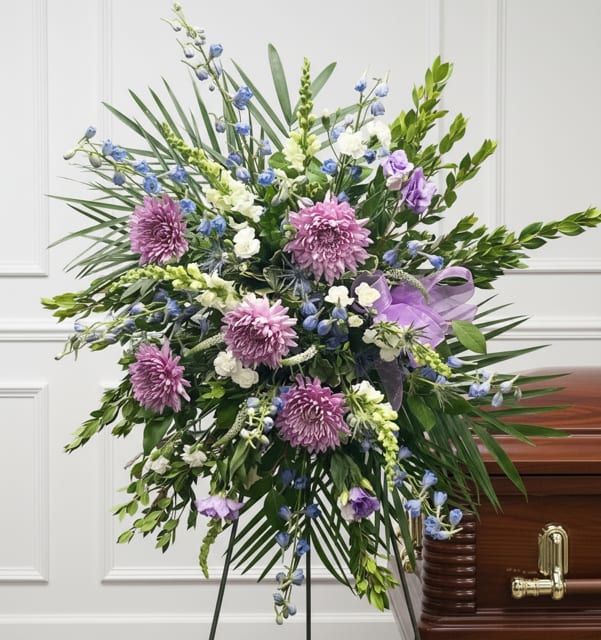 Large funeral spray with purple and blue flowers on a stand beside a casket