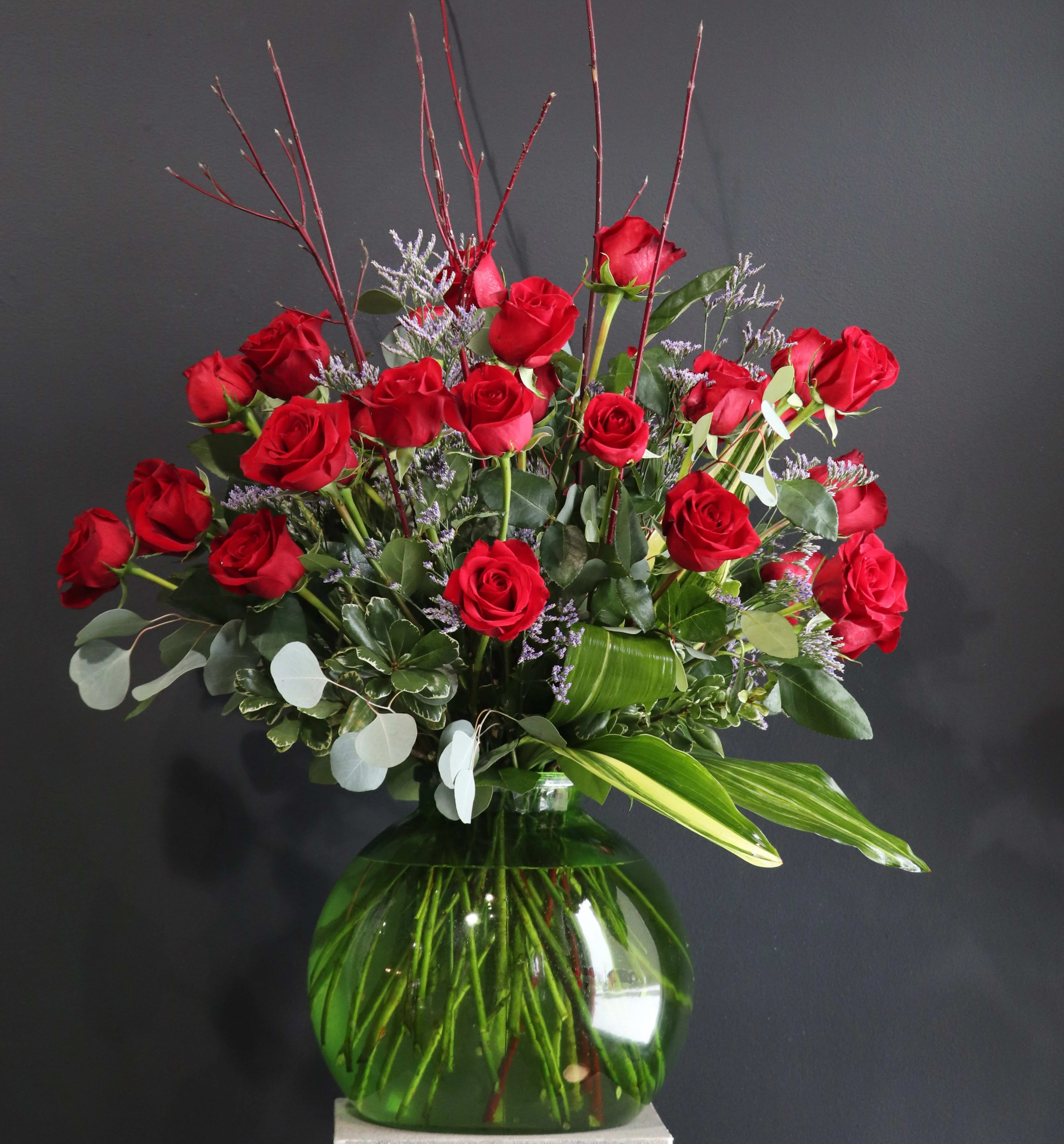 Red roses arranged in a green glass vase with purple filler and branches