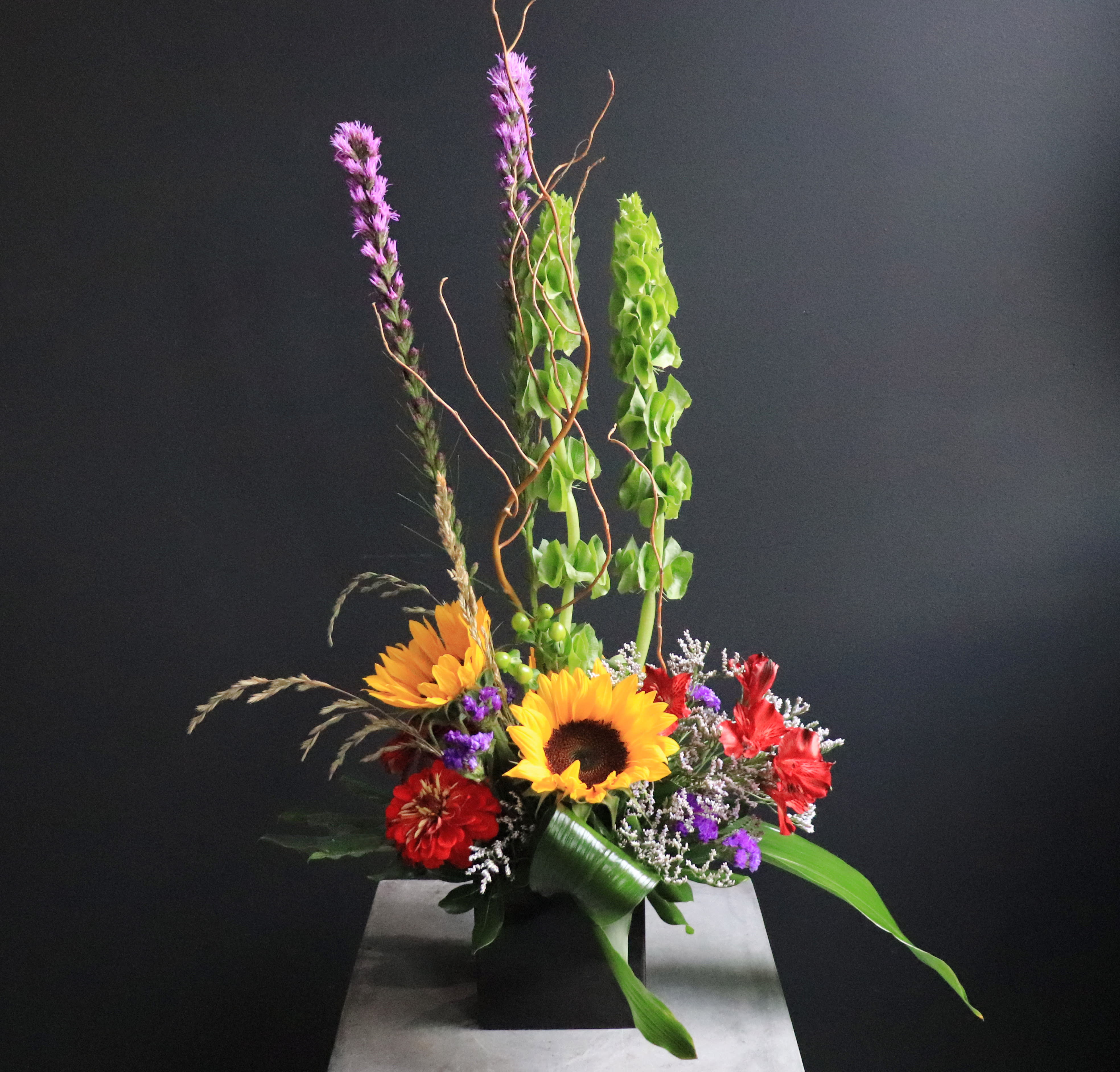 Tall mixed bouquet with sunflowers, red blooms, and green accents in a black vase