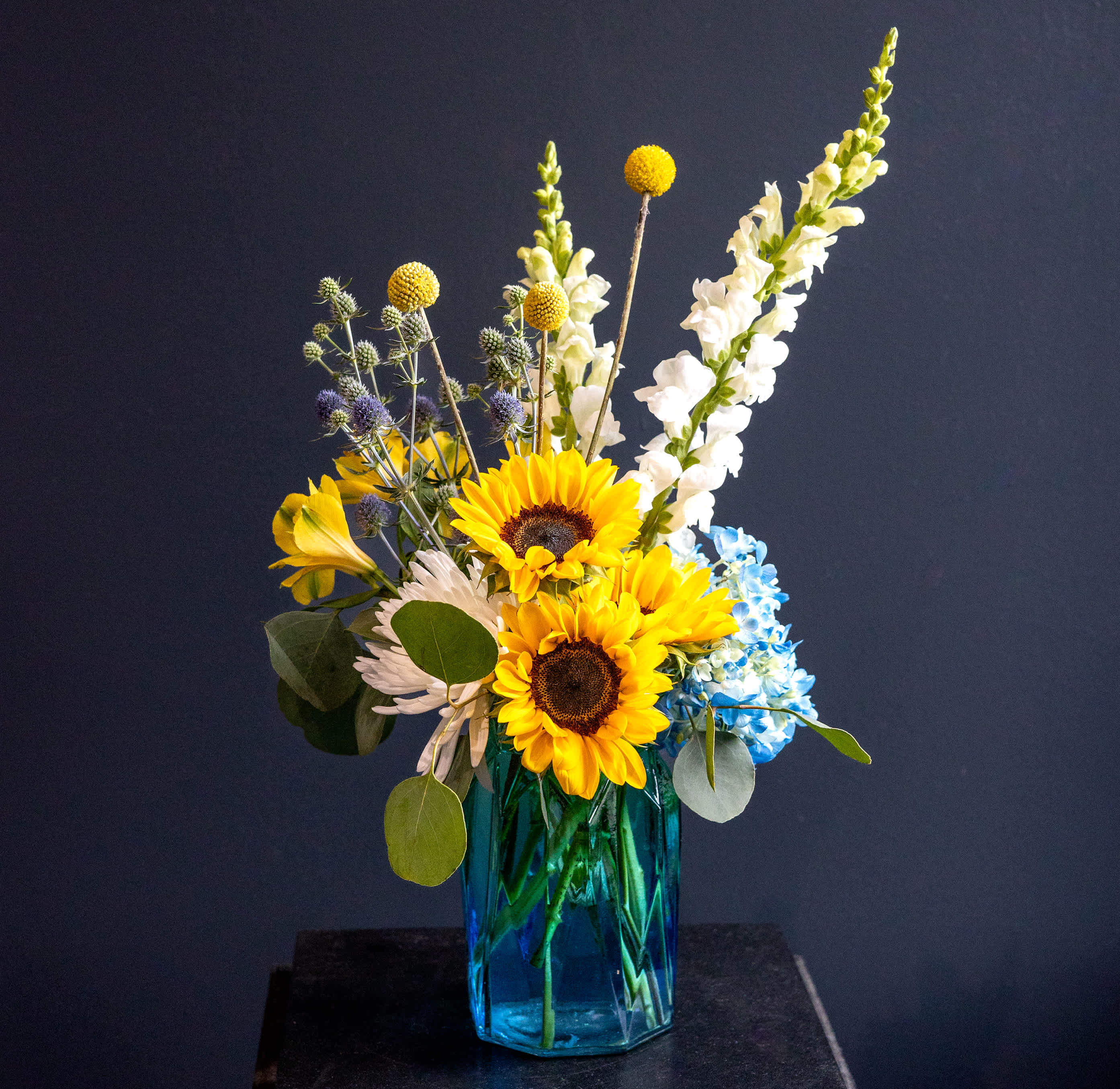 Sunflowers, white snapdragons, and blue hydrangeas in a blue glass vase
