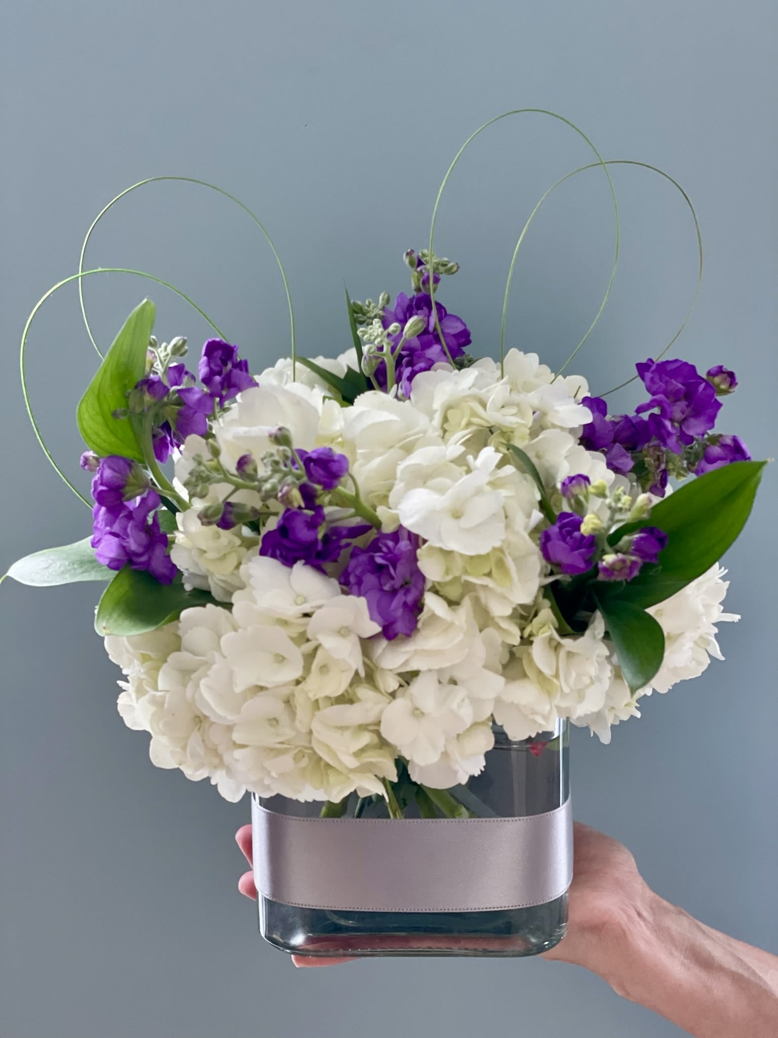 White hydrangeas and purple stock in a glass vase with looping wire accents