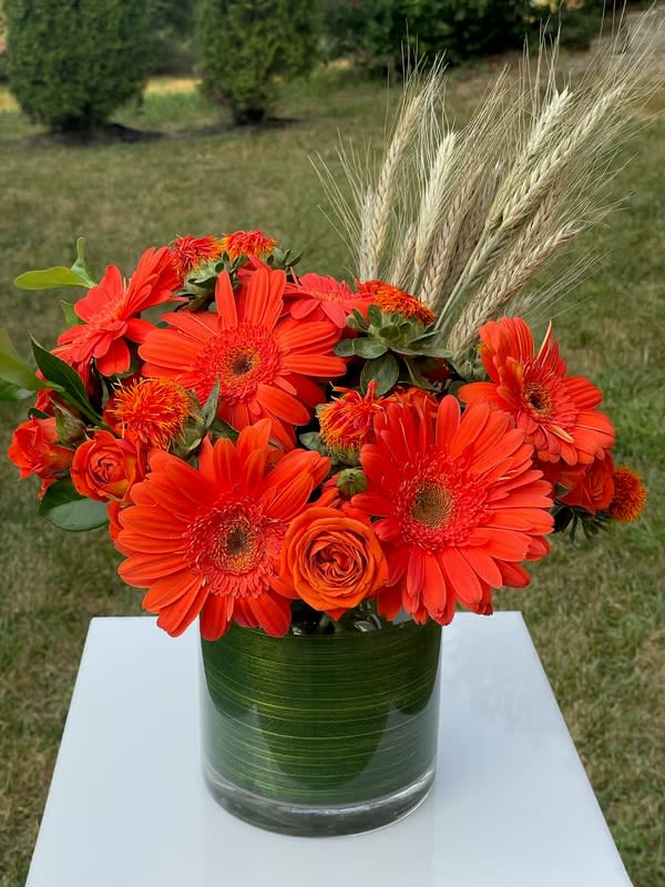 Orange gerbera daisies and roses in a glass vase with wheat stems