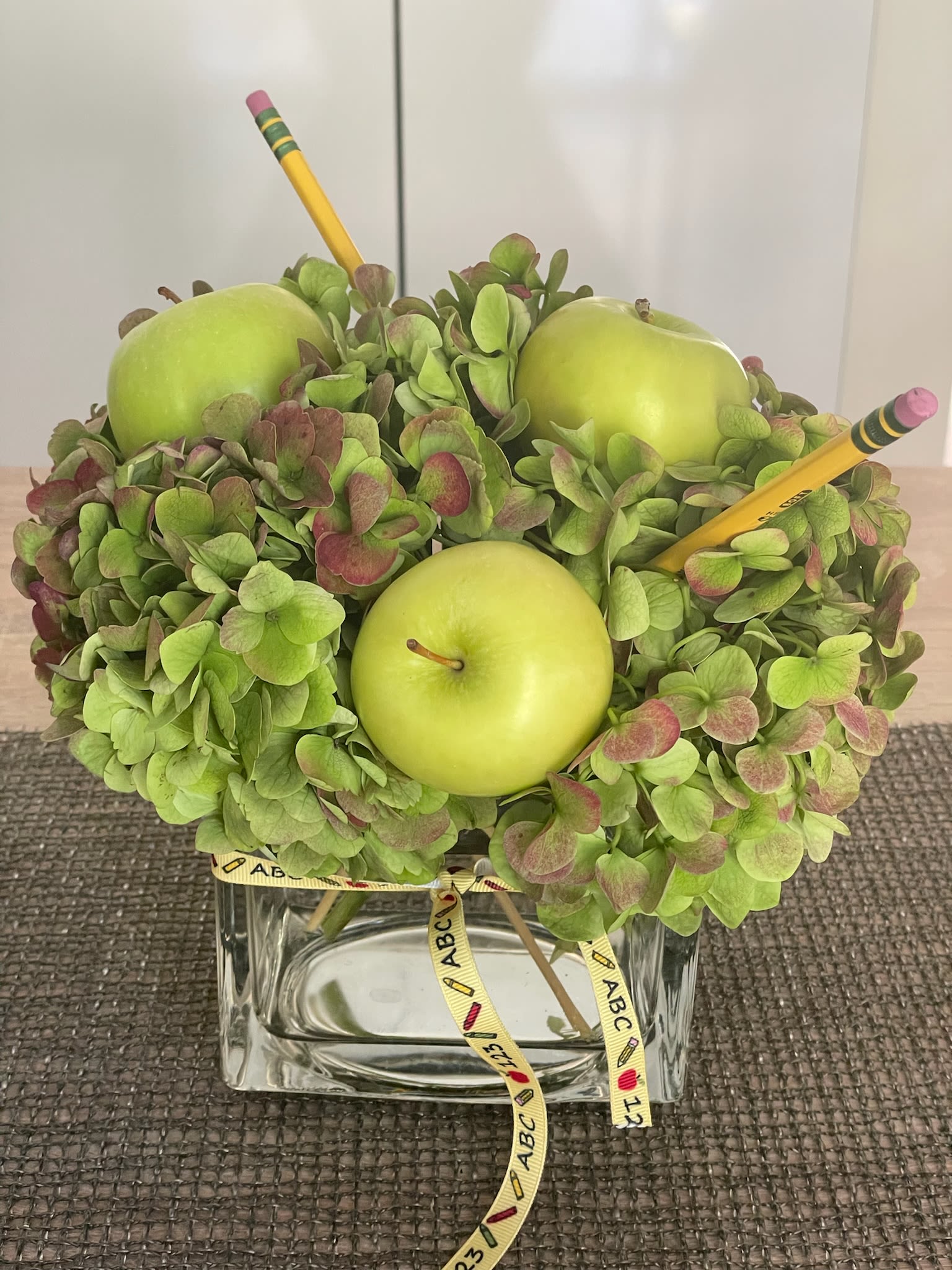 Green hydrangea arrangement with apples and pencils in a glass vase