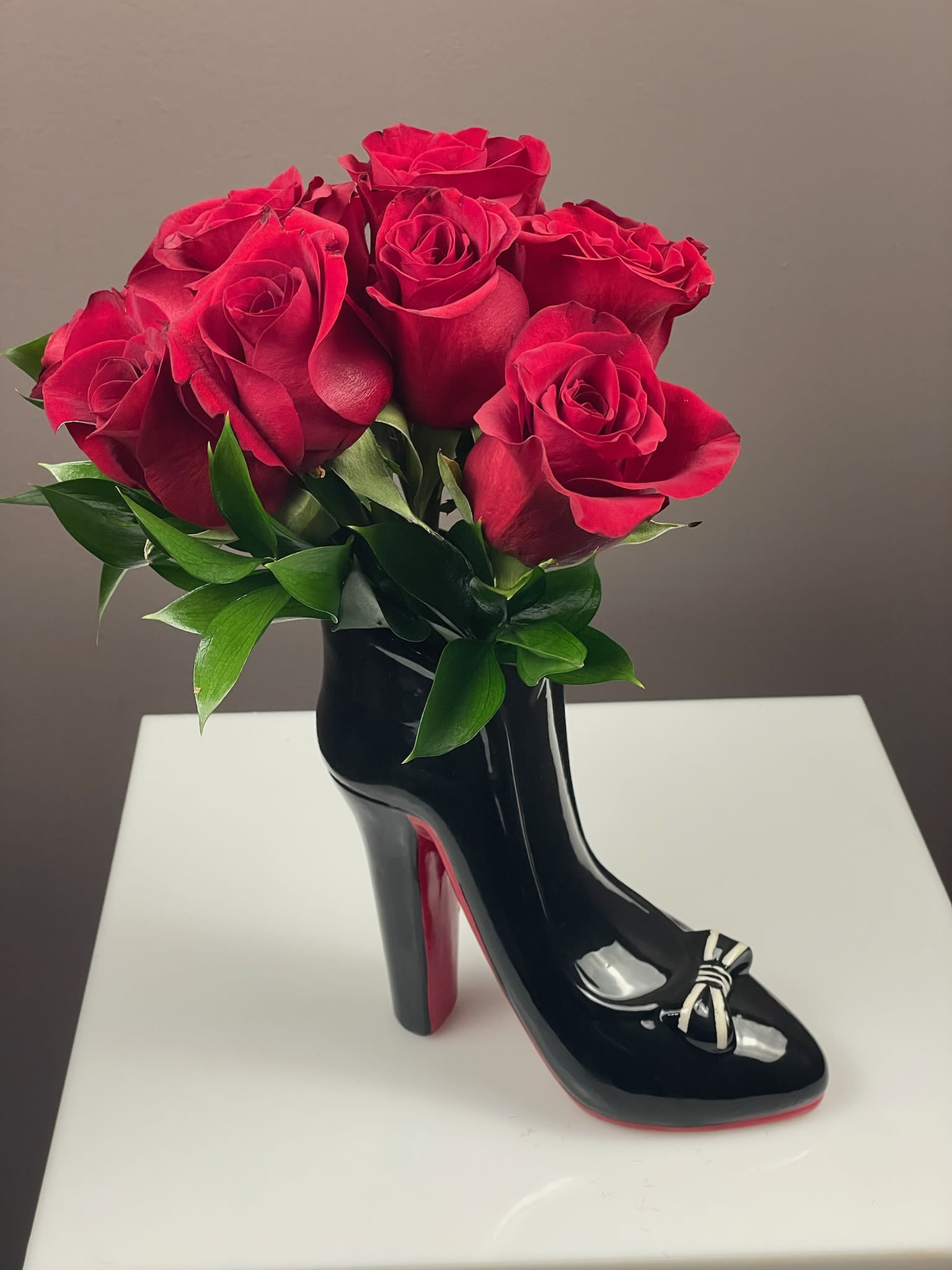 Stiletto Statement - Step into style with our most requested floral arrangement! This chic, high-fashion statement piece from Blooming Bouquet is the perfect gift for the trendsetter in your life or a sophisticated addition to your own home decor.  This unique design features a premium selection of roses nestled in a sleek high-heel vase. The glossy black finish, accented with a classic bow and a bold red sole, pays homage to iconic luxury footwear.  Product Details:  Dimensions: Stands approximately 12 inches high and 8 inches wide, making it the perfect size for a vanity, office desk, or side table. Floral Choice: Available in your choice of Classic Crimson Red or Elegant Creamy White roses. Design: A lush, rounded bouquet accented with fresh, deep green ruscus foliage for a polished look. The Vase: A reusable, high-gloss stiletto that serves as a beautiful keepsake long after the flowers have bloomed. Special Touches: The red rose option includes a plush heart pick, making it an ideal choice for anniversaries, birthdays, or Valentine's Day.