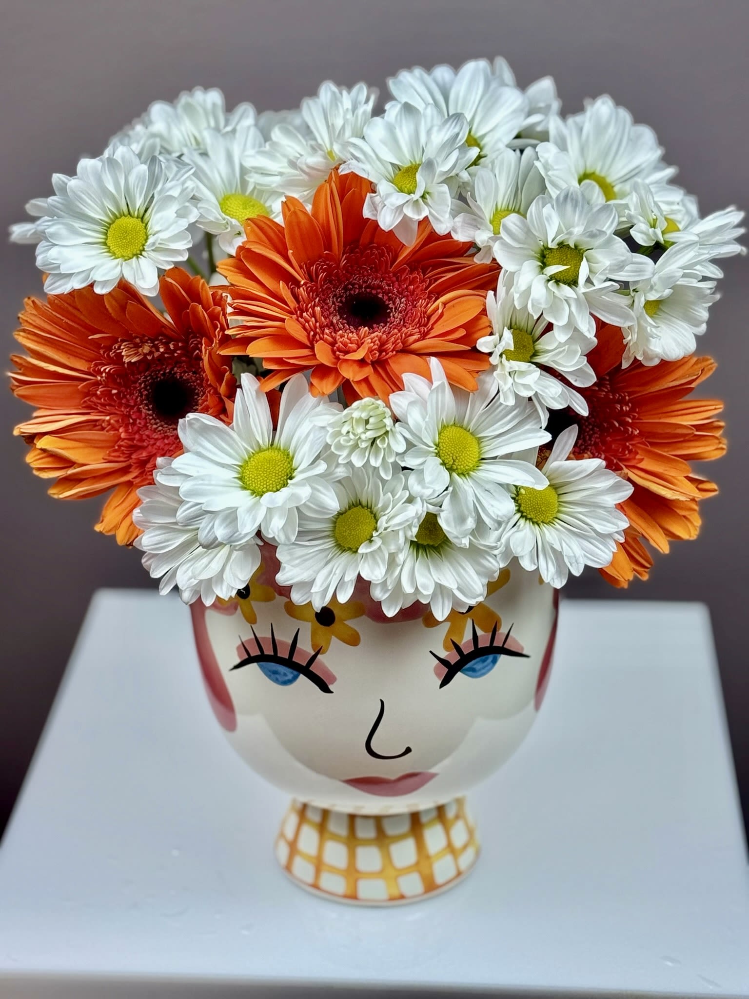 Whimsical Wink - This cheerful arrangement is designed to bring a playful burst of personality and color to any space. Featuring a vibrant mix of bold orange Gerbera daisies and crisp white chrysanthemums, this bouquet is perfectly balanced to offer both energy and elegance.  The flowers are artfully nestled in a unique, hand-painted ceramic "face" vase that adds a touch of modern charm and artistic flair. Whether you’re looking to brighten a home office or send a thoughtful gift that stands out, this arrangement is sure to spark a smile.  Product Details:  Vase: Decorative "Face" ceramic pedestal vase with whimsical hand-painted details.  Dimensions: Approximately 12 inches high by 11 inches wide.  Best For: Birthdays, "thinking of you" gifts, or adding a pop of color to your own decor.  Bring home a bit of blooming joy today! You can order this unique piece directly through our website for local delivery.
