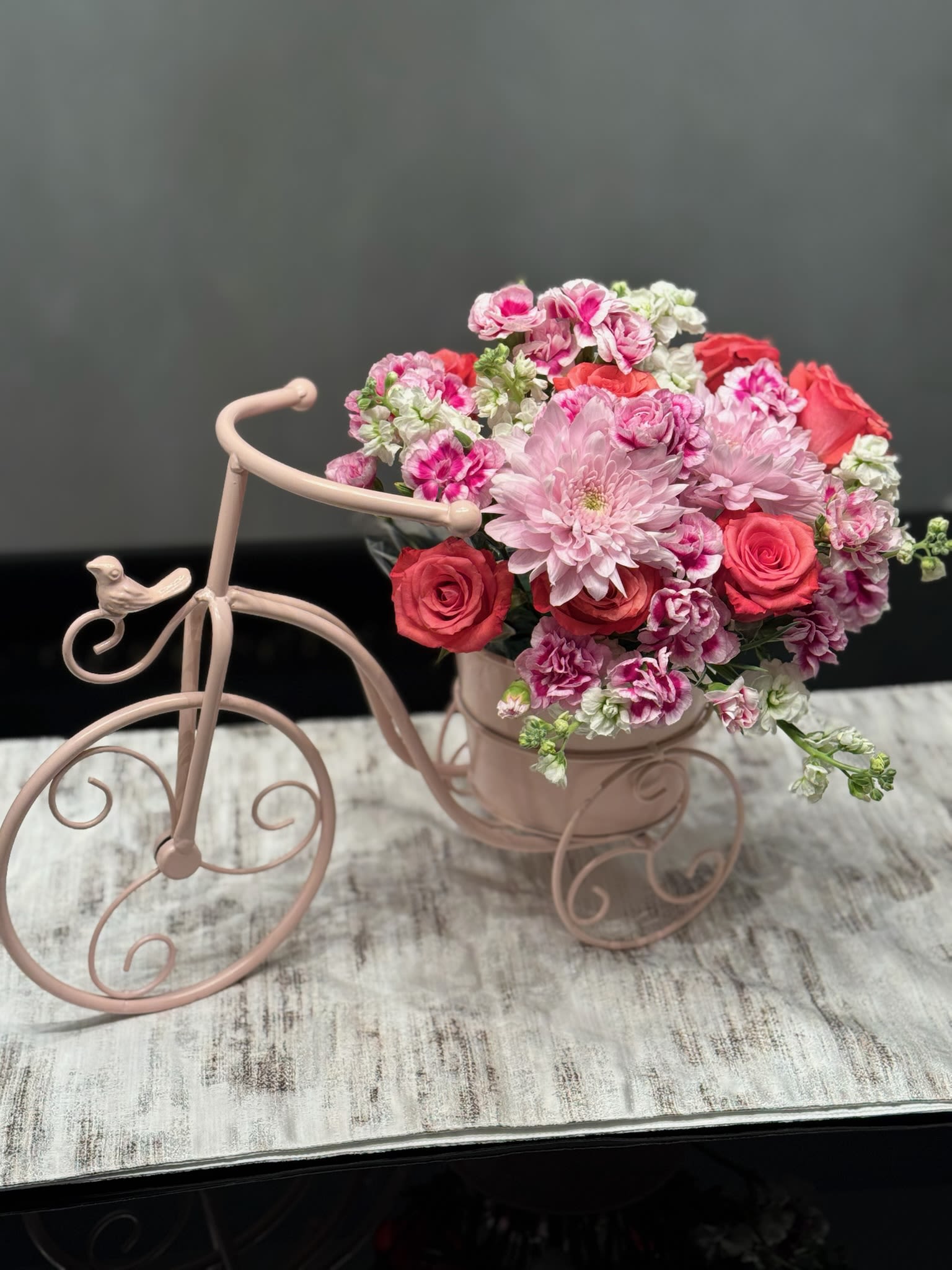 Whimsical Bicycle Flower Arrangement - This charming, vintage-inspired arrangement is a masterclass in texture and color, blending romantic pinks and deep corals into a lush, rounded display. The dense canopy of ruffled petals and star-shaped blooms creates a soft, layered silhouette that radiates warmth and elegance from every angle.  The flowers are artfully nestled within a whimsical, powder-pink metal bicycle planter. With its delicate scrollwork wheels and a tiny bird perched gracefully on the handlebars, the container adds a playful, storytelling element to the design. It’s a perfect balance of sophisticated floral artistry and sweet, keepsake charm—ideal for a nursery, a kitchen island, or as a thoughtful gift that remains a beautiful decorative piece long after the flowers have finished their show.  Height 14 in. Width  16 in.