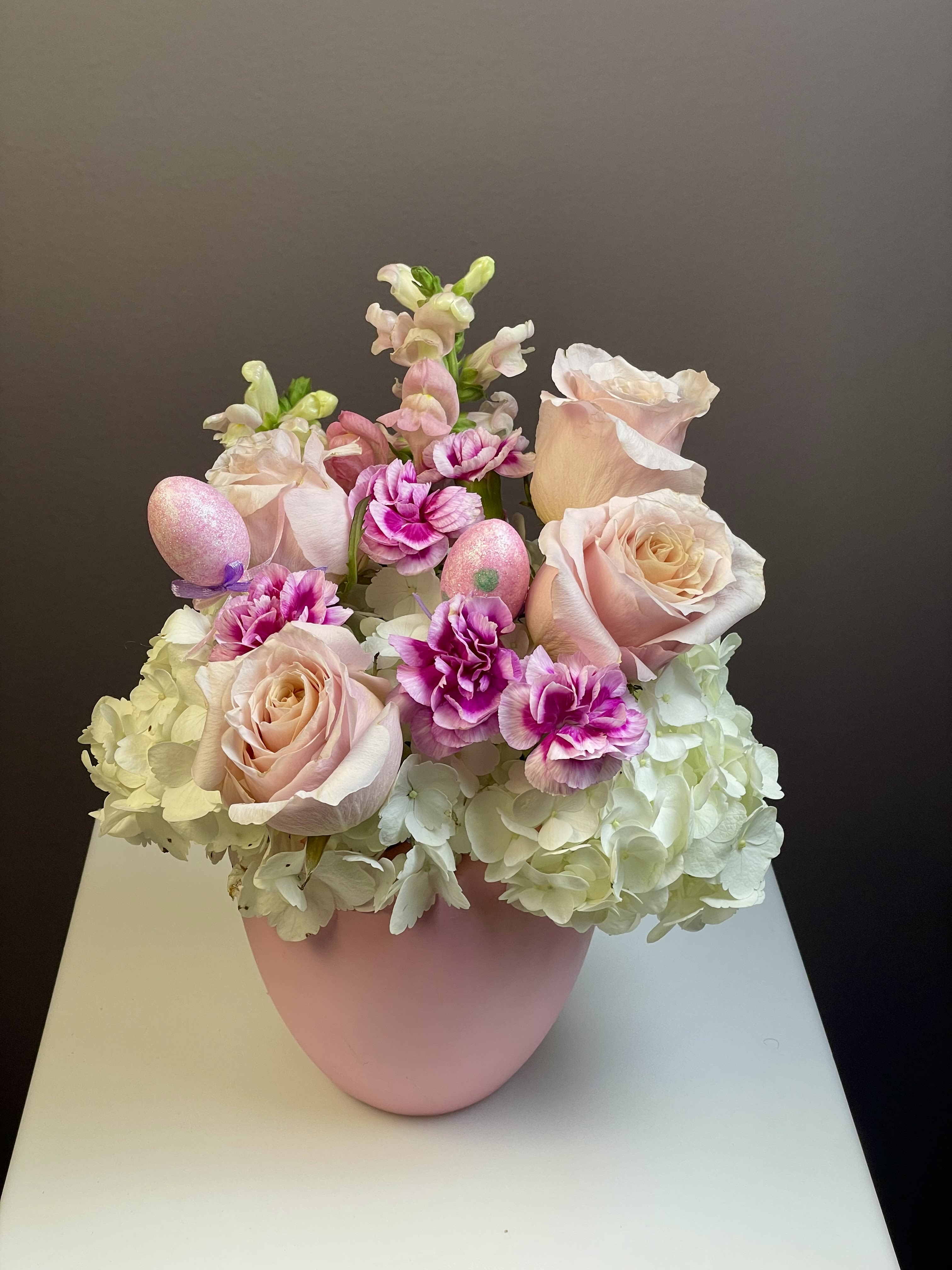 Some-Bunny Special - Hop into spring with our Some-Bunny Special arrangement! This playful Easter design is a delightful mix of soft textures and festive accents, perfectly curated to brighten any holiday table or surprise a loved one.  The Design   This arrangement features a lush base of snowy white hydrangea, topped with premium long-stemmed blush roses and ruffled bicolored carnations in vibrant pink. For that extra festive touch, we’ve nestled     sparkling pink glitter eggs among the blooms, all gathered in a sleek, matte pink ceramic vase.  Choose Your Look - Customized for your style, this arrangement is available in two versions:   With Bunny Ears: Includes whimsical, iridescent pink "bunny ears" for a playful, family-friendly centerpiece.    Classic Floral: A sophisticated, egg-accented spring bouquet without the ears—perfect for a modern Easter brunch.