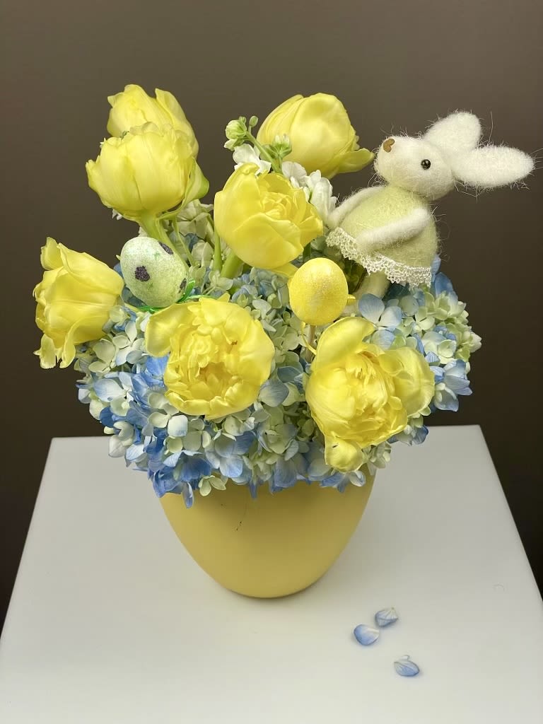 Sunny Bunny - Bring the joy of spring into your home with our Sunny Bunny Easter arrangement! This vibrant design features a base of cloud-like blue hydrangea topped with cheerful yellow tulips and seasonal accents. For a truly festive touch, it includes a charming felt bunny pick and speckled Easter eggs, all nestled in a sunny yellow ceramic vase.  Customizable Beauty - Tailor this arrangement to your recipient's favorite flower:   Tulip Version (Pictured): Features bold, bright yellow tulips for a classic spring look.   Rose Version: Can be crafted with premium yellow roses for a touch of timeless elegance.  Product Details   Perfectly Sized: Standing 11 inches tall and 10 inches wide, it is the ideal size for a breakfast nook, side table, or as a bright holiday centerpiece.   Festive Accents: Includes a handcrafted felt bunny and decorative Easter eggs for a whimsical holiday feel.