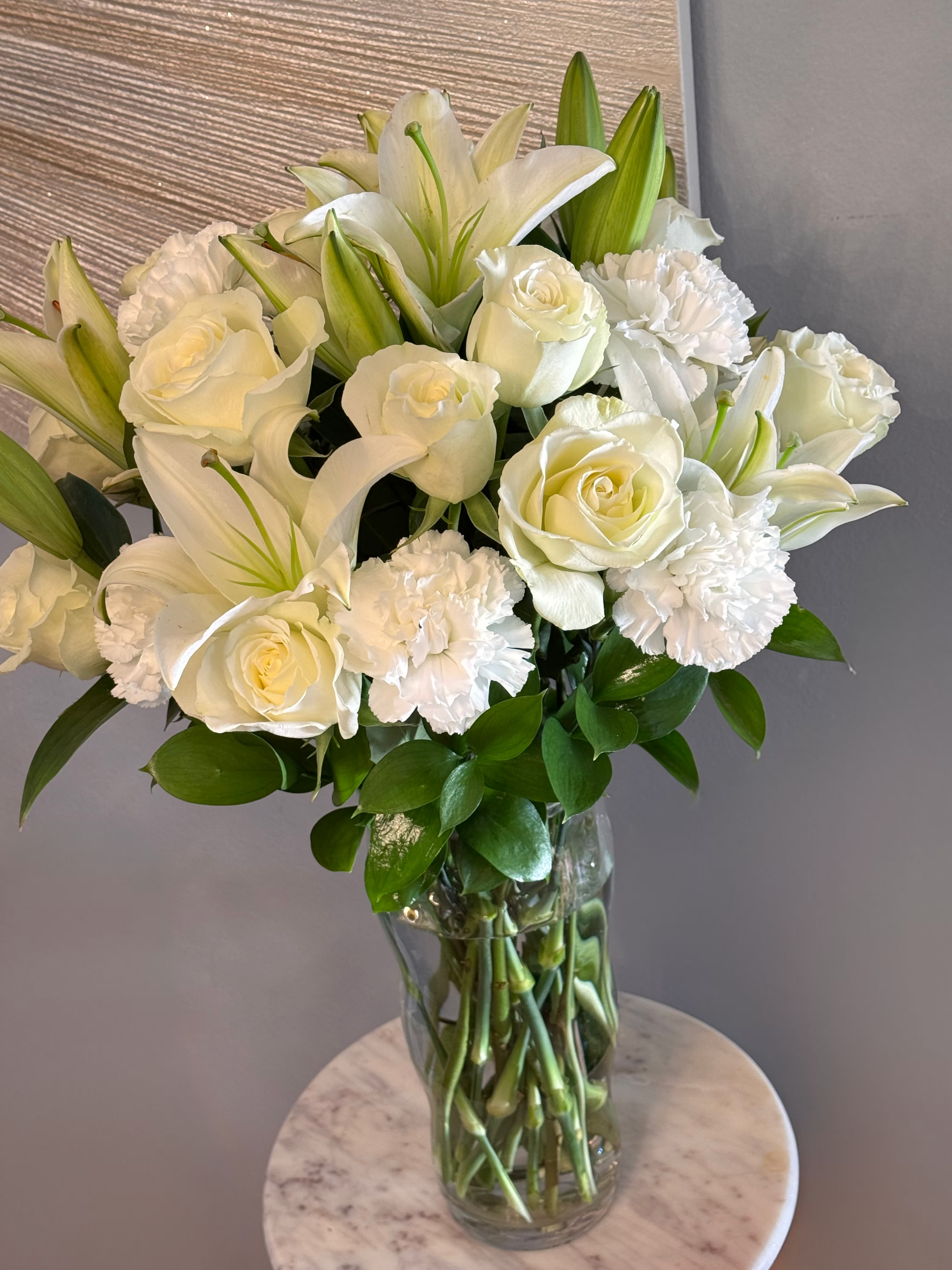 Timeless Ivory Elegance - This timeless floral arrangement features a luxurious blend of pure white roses, graceful lilies, and soft white carnations, accented with lush green foliage. Designed in a clear glass vase, the stems are beautifully displayed, creating a clean and elegant look from every angle. Perfect for sympathy, celebrations, or refined home décor, this arrangement conveys serenity, grace, and lasting beauty.