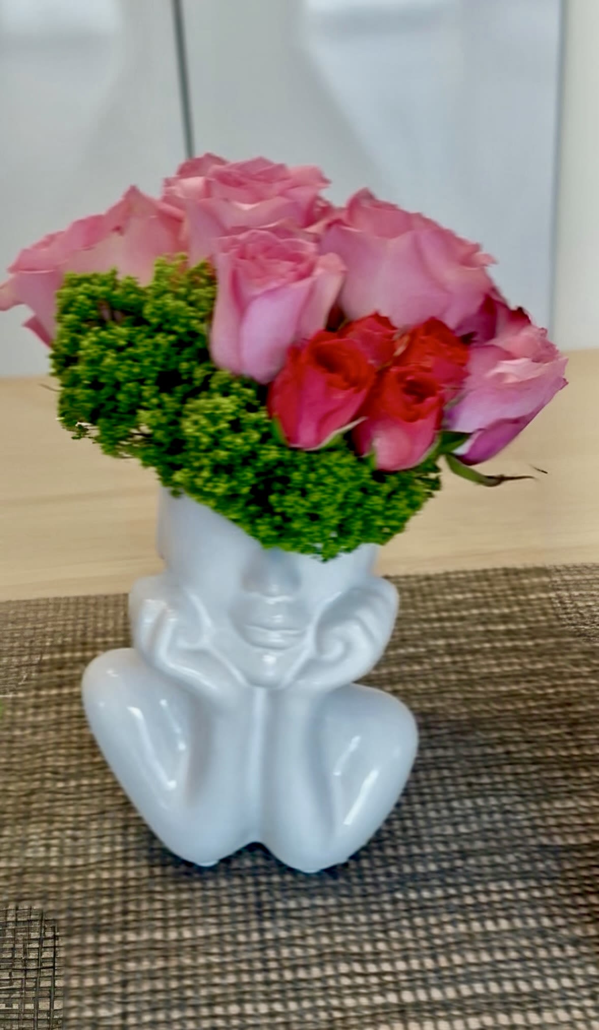 Grace Bouquet - Our "Grace Bouquet" is a tribute to the boundless strength and nurturing spirit of women everywhere. Encased in a vase sculpted in the graceful form of a woman's torso, with her head tenderly cradled in her palms, this arrangement exudes elegance and maternal love. Bright pink roses, symbolizing admiration and affection, are complemented by vibrant green dianthus, signifying growth and harmony. Together, they create a harmonious blend of beauty and vitality, reflecting the multifaceted nature of motherhood. This arrangement is a heartfelt expression of gratitude and appreciation for the endless love and support that mothers provide.  Height 12 in Width 8 in