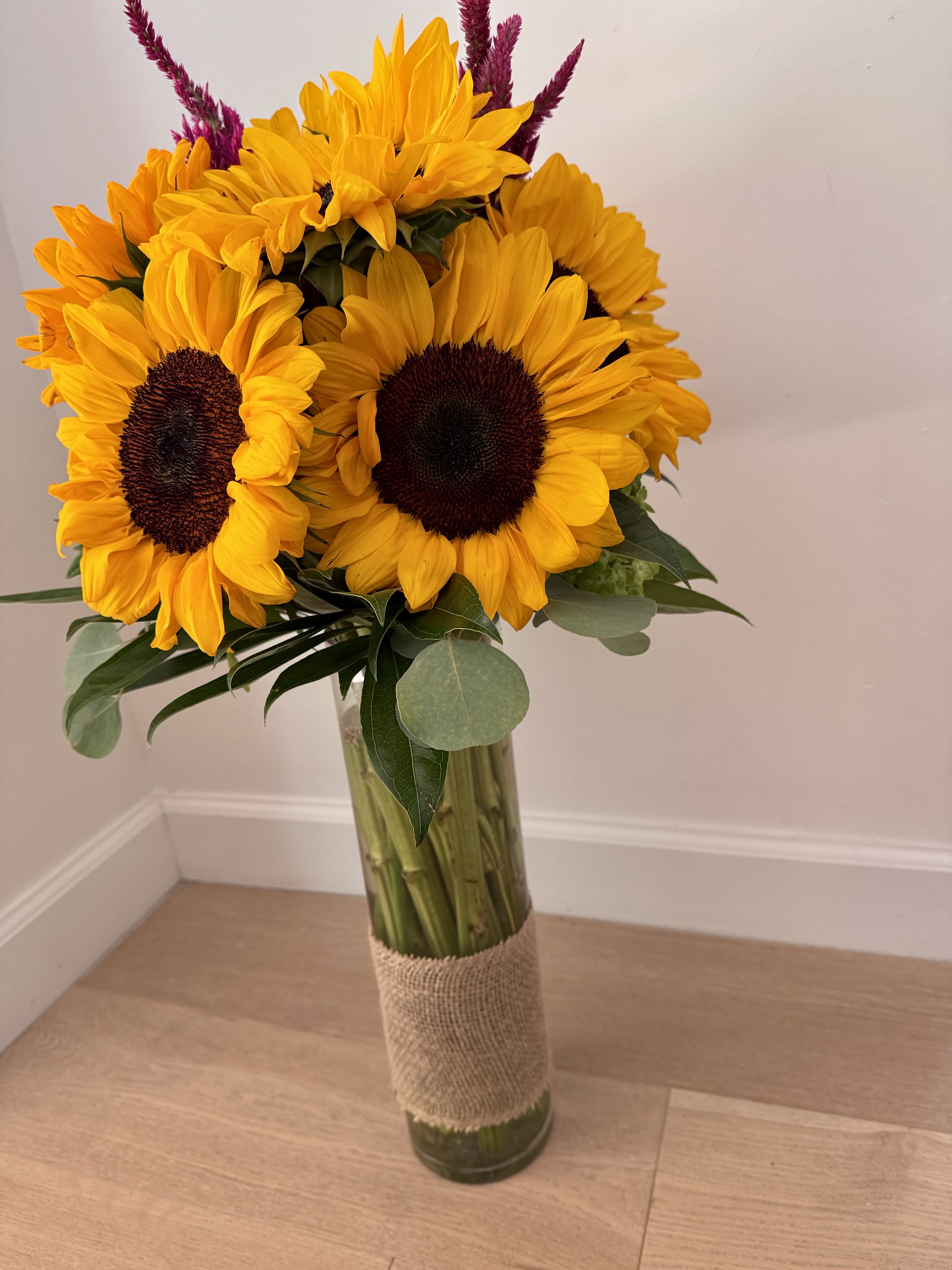 Sunflower Cheer Arrangement - A radiant display of sunflowers designed to brighten any space. This arrangement features large, golden blooms paired with vivid magenta accents and lush greenery, creating a lively, uplifting composition. The flowers are styled in a clear glass vase wrapped with natural burlap for a warm, rustic finish. Perfect for celebrations, thank‑you gifts, or simply bringing sunshine indoors.  Key features:  Sunflower blooms — bold, golden petals with rich centers Magenta accents — added color contrast for visual depth Rustic burlap wrap — a natural, handcrafted touch Clear glass vase — showcases fresh stems and clean design Long‑lasting freshness — ideal for home or office décor