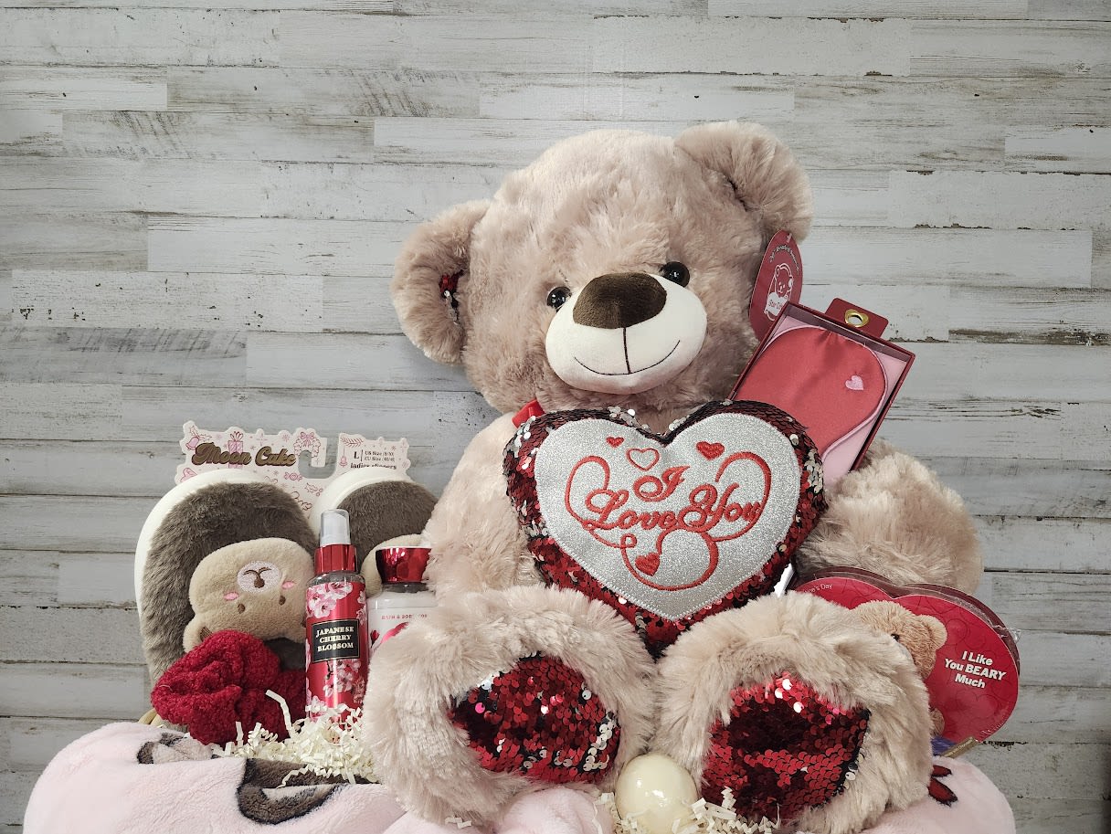Large teddy bear holding a heart pillow with assorted gift items