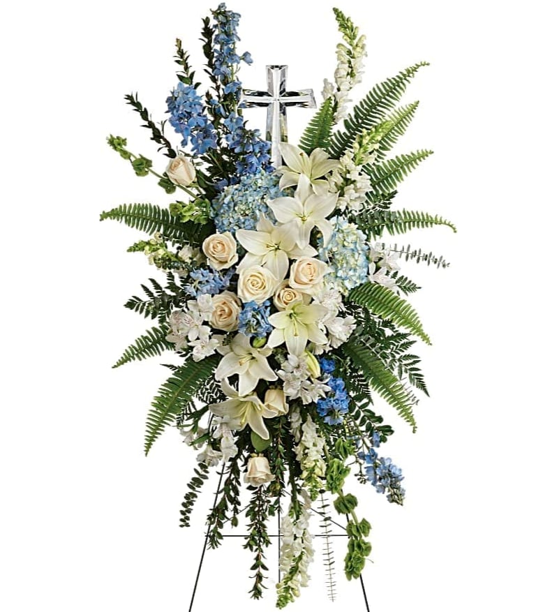 Standing funeral spray with white lilies, cream roses, and blue flowers on a cross stand