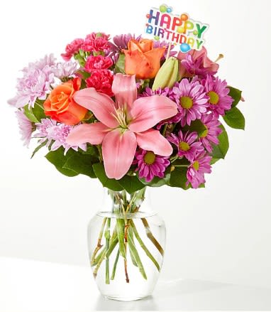 Mixed pink and orange flowers in a clear glass vase with a birthday topper