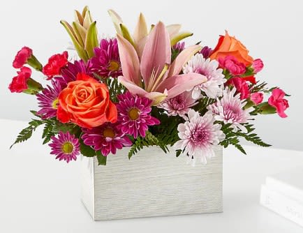 Rectangular arrangement of pink lilies, orange roses, and purple and white daisies in a white box.