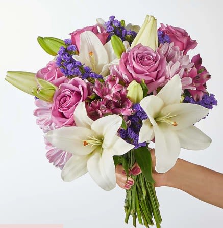 Hand-tied bouquet of white lilies, pink roses, and purple filler flowers held against a light background