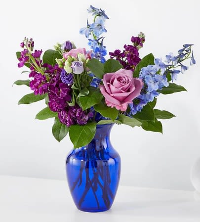 Purple, blue, and pink flower arrangement with roses and delphinium in a cobalt blue glass vase