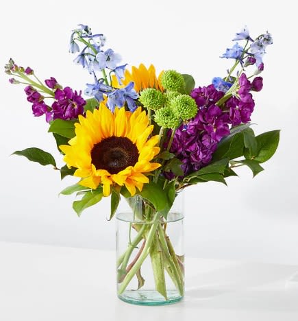 Bright sunflower, purple stock, blue delphinium, and green mums in a clear glass vase.