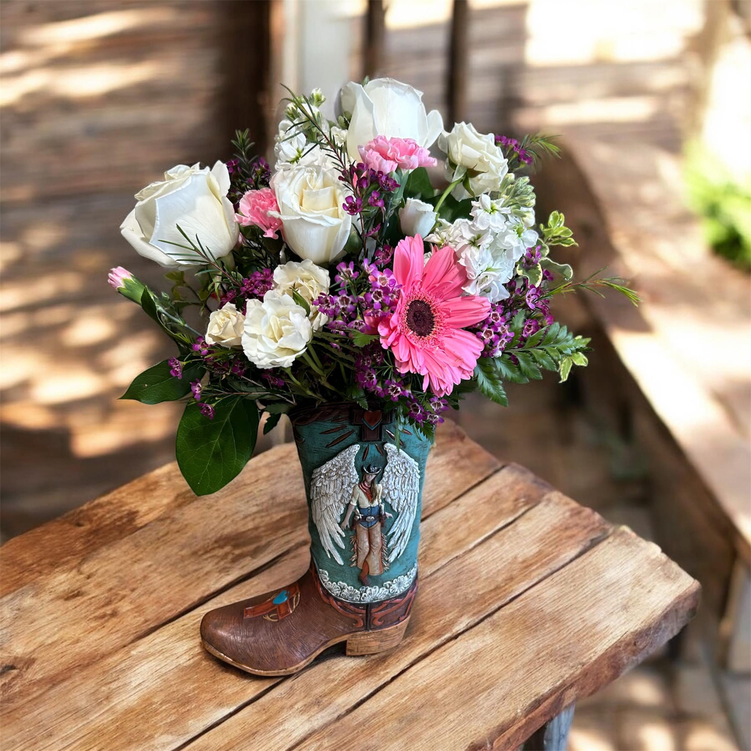 Blushing Cowgirl Boot - This charming Western-inspired arrangement is designed in a decorative cowboy boot vase, bringing together rustic flair and soft elegance. It features a beautiful mix of creamy white roses, soft pink carnations, a vibrant pink gerbera daisy, and delicate accent blooms in shades of purple and white. Lush greenery adds fullness and texture, creating a balanced and eye-catching display.  Perfect as a gift or home décor piece, the “Blushing Cowgirl Boot” blends country charm with romantic tones—ideal for birthdays, thank-you gifts, or just because.