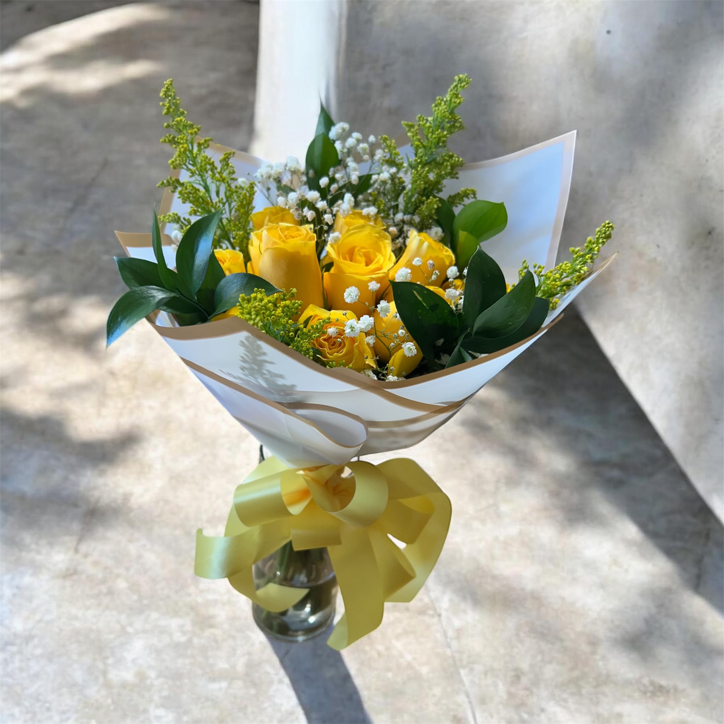 Yellow Bliss Wrap - This bright and cheerful hand-wrapped bouquet features 6 vibrant yellow roses accented with delicate solidago and fresh greenery. The soft filler adds a light, airy texture while enhancing the rich golden tones of the roses. Carefully wrapped for a clean and elegant finish, this bouquet is simple yet full of warmth.  Perfect for celebrating friendship, sending well wishes, or just because, the “Golden Glow Half Dozen” is a beautiful expression of happiness and positivity.