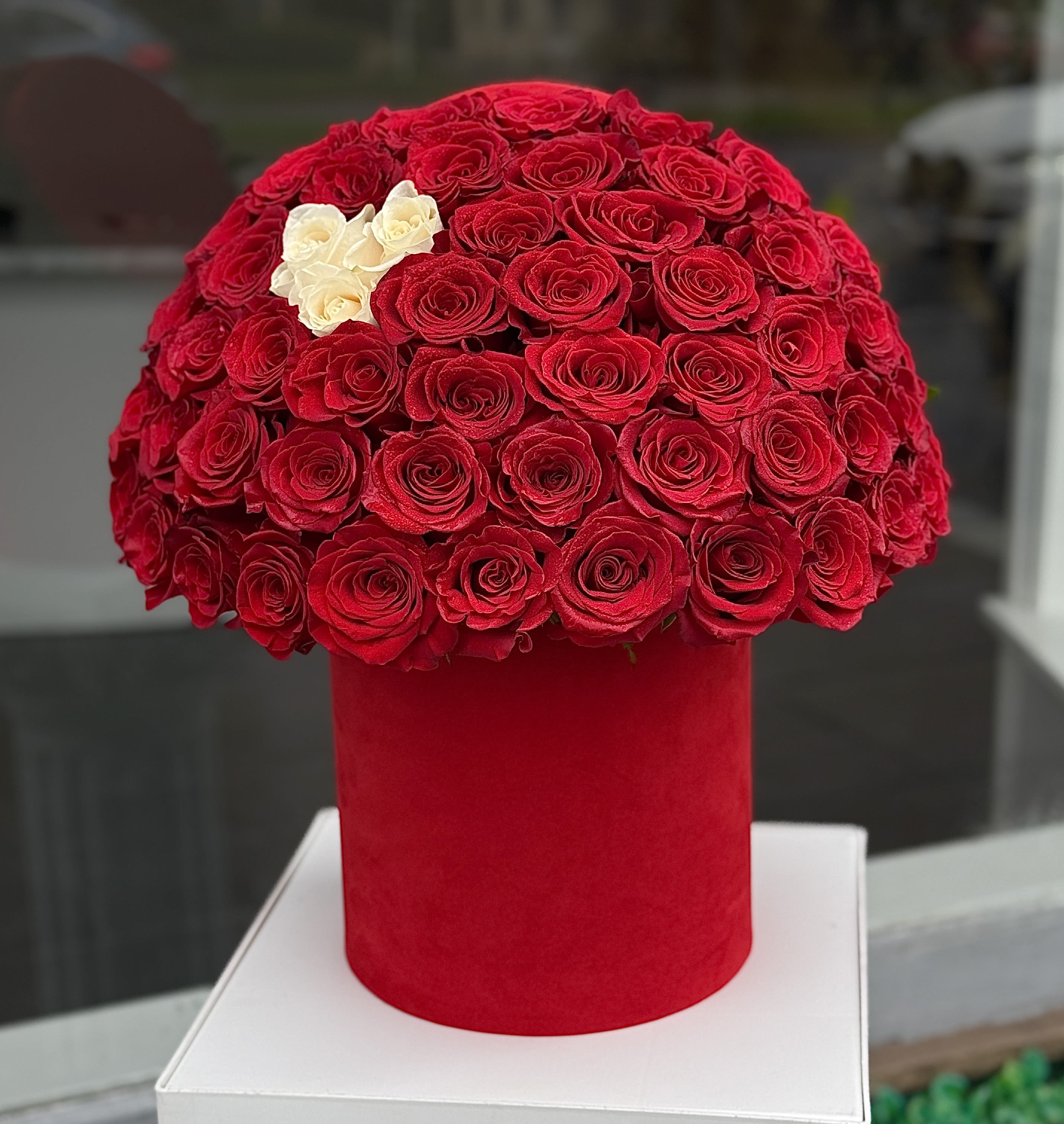 Red rose arrangement in a red cylindrical box with a few white roses