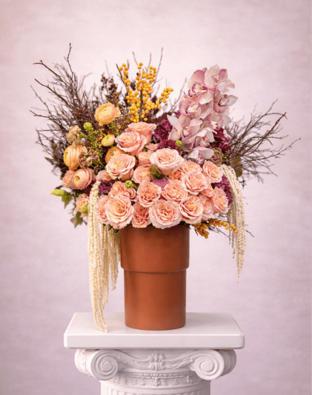 Tall arrangement of peach roses, ranunculus, orchids, and yellow berries in a brown vase on a white pedestal.