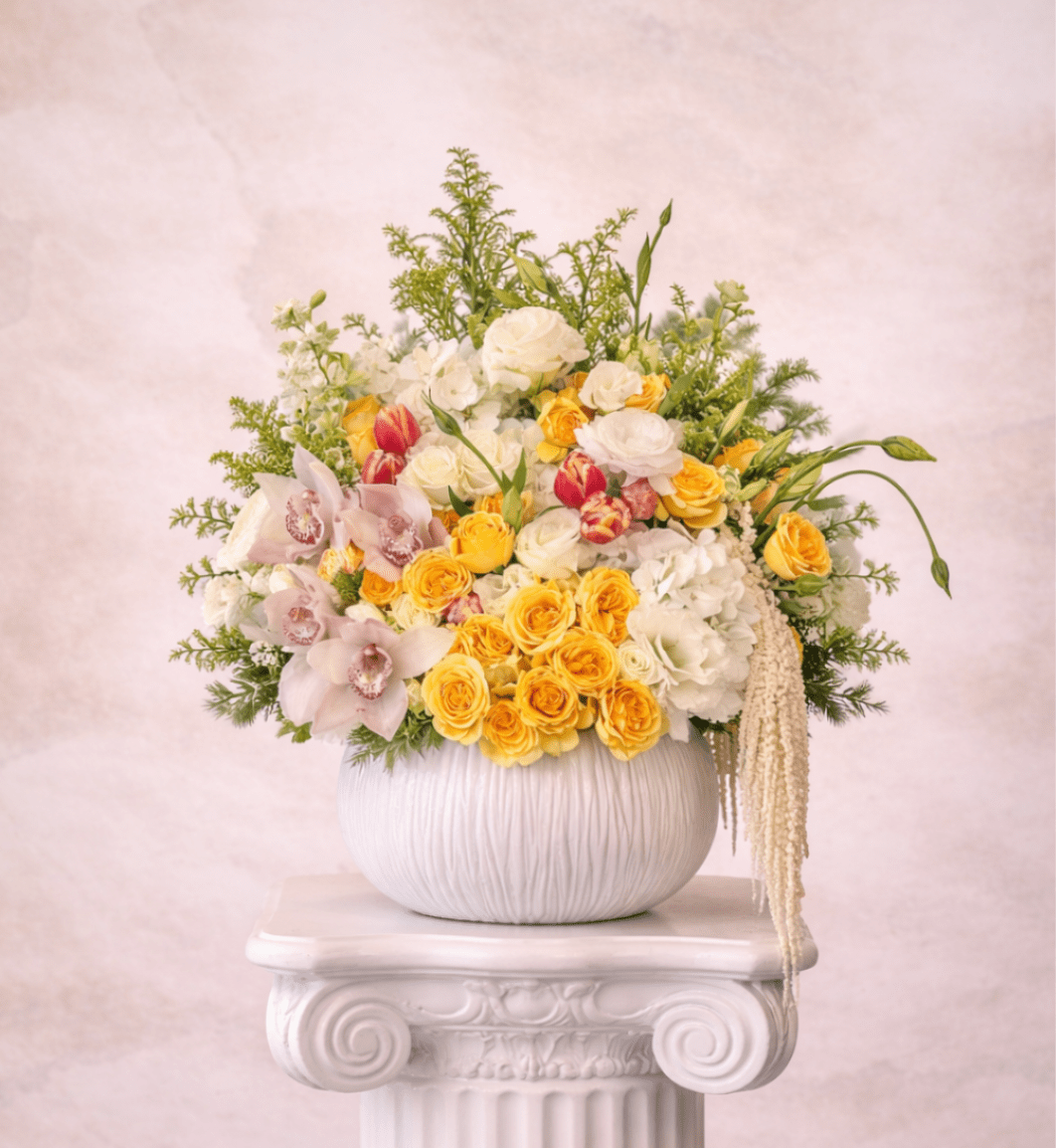 Yellow and white floral arrangement with roses, orchids, and tulips in a round white vase on a pedestal.
