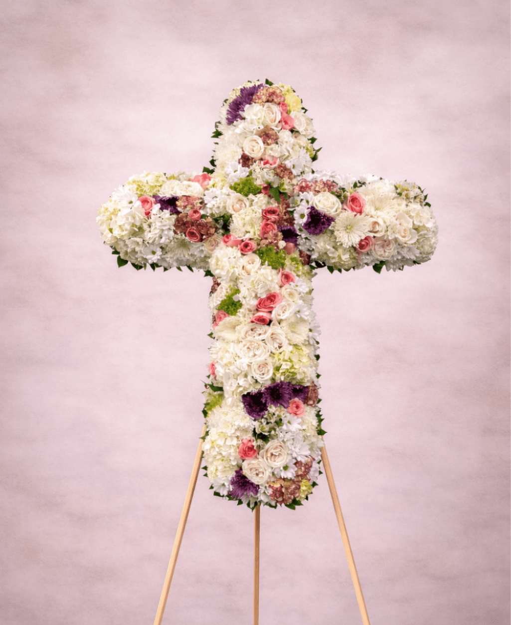 Standing floral cross of white blooms with pink roses and purple accents on a wooden easel