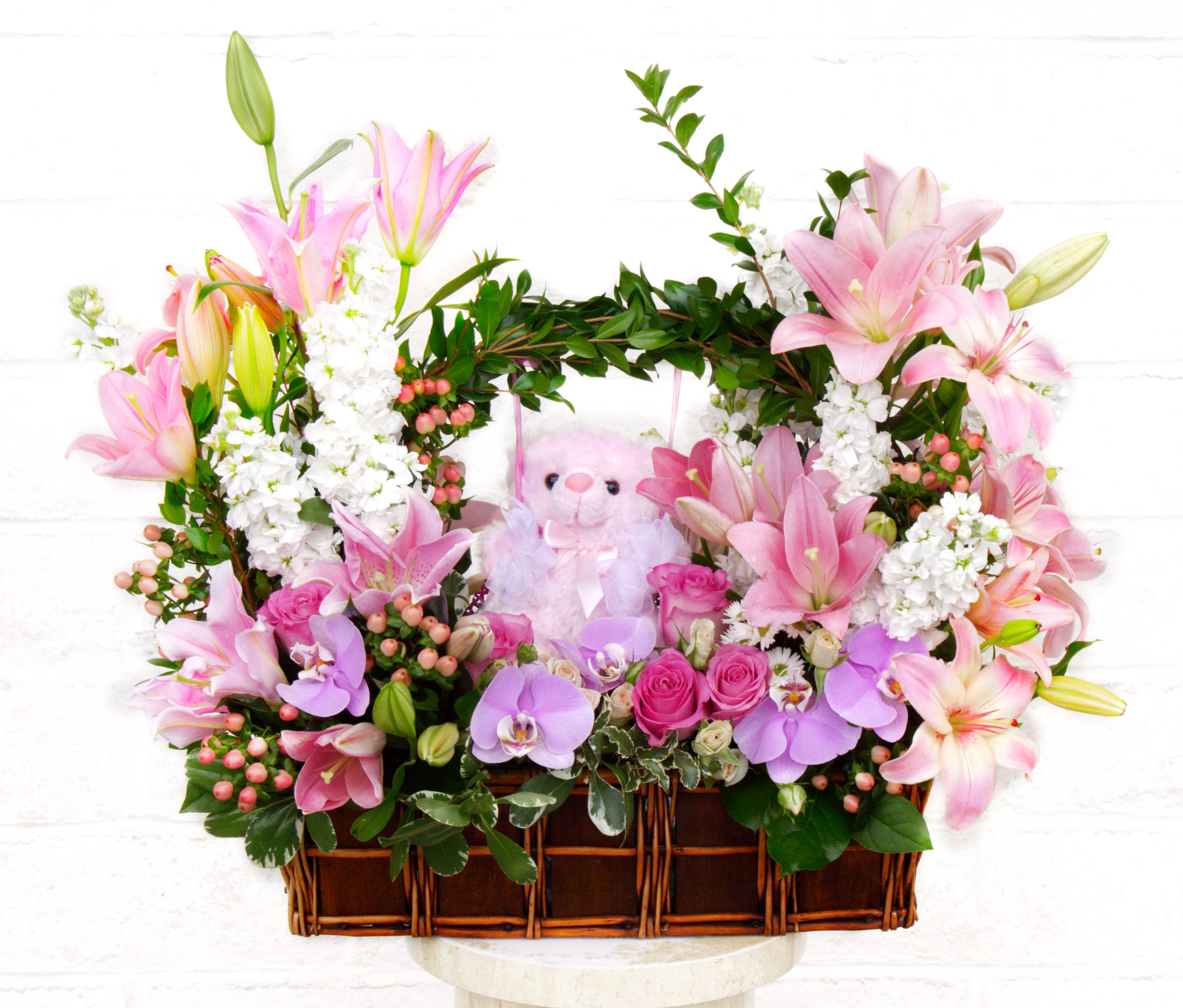 Pink lilies and orchids arranged in a wicker basket with a teddy bear