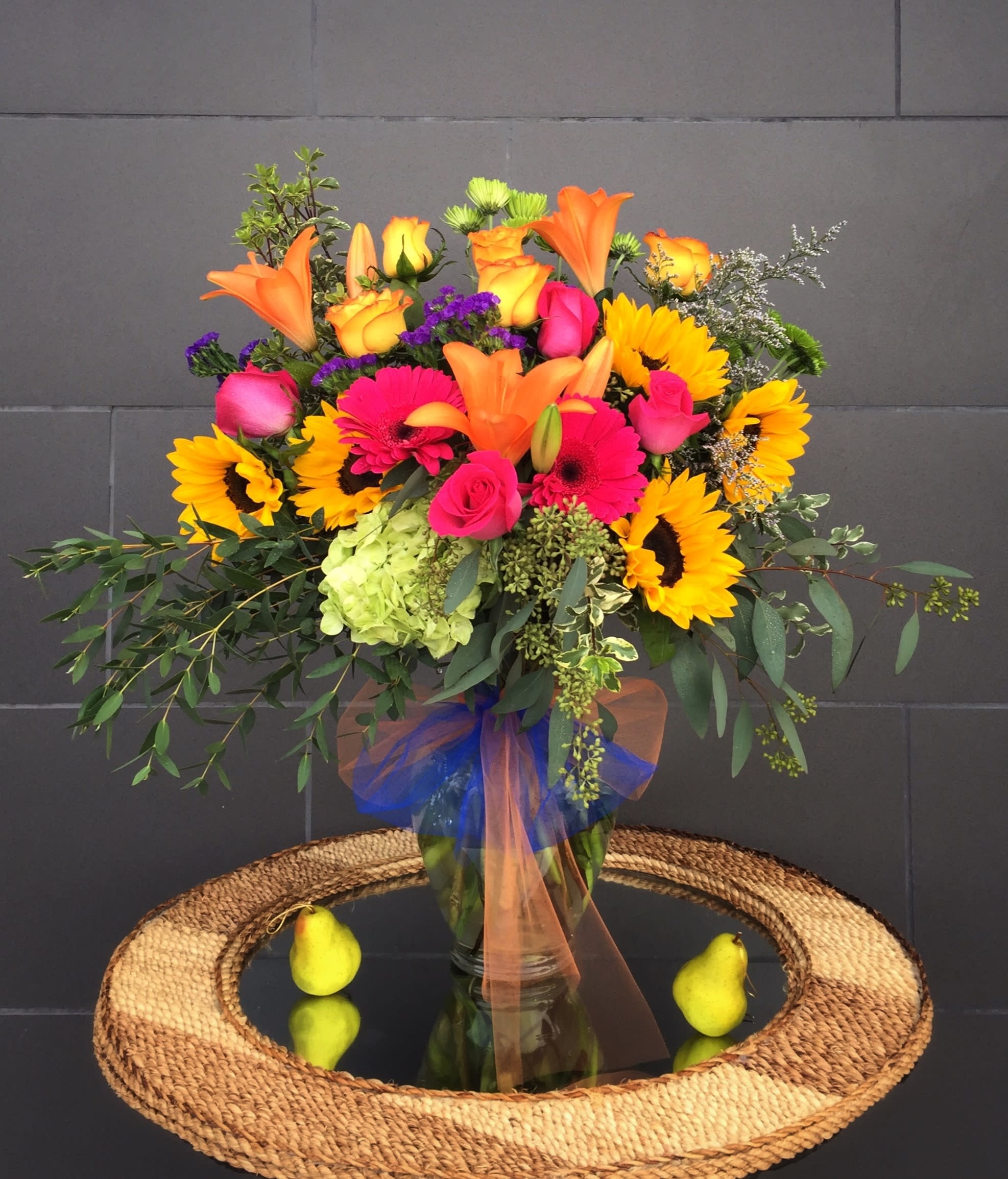 Bright mixed bouquet with sunflowers, roses, lilies, and gerbera daisies in a glass vase.