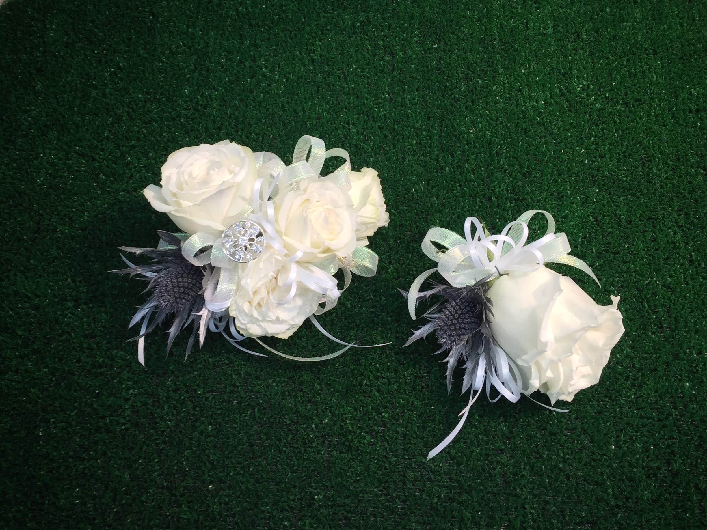 Two white rose corsages with ribbon and dark feather accents on green turf