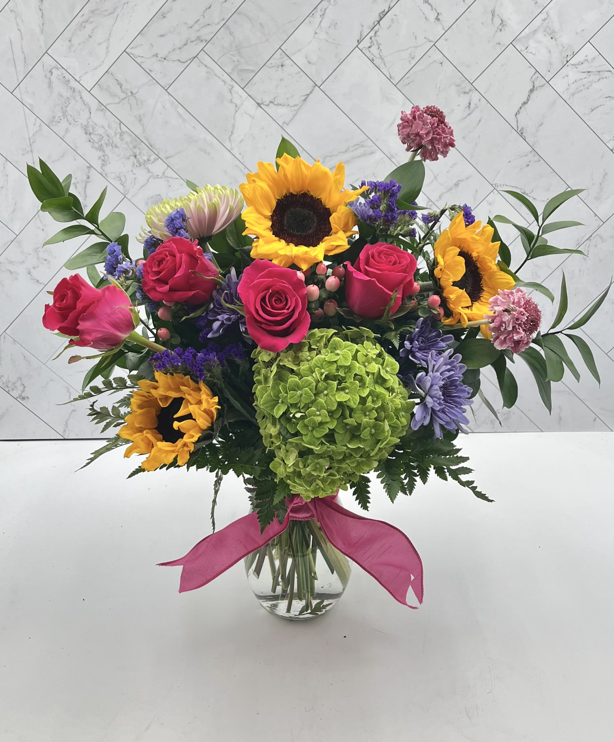 FANCY FIESTA - An arrangement for any celebration. Features, sunflowers, roses, hydrangeas, scabiosa and more.  Arranges in a tall vase  