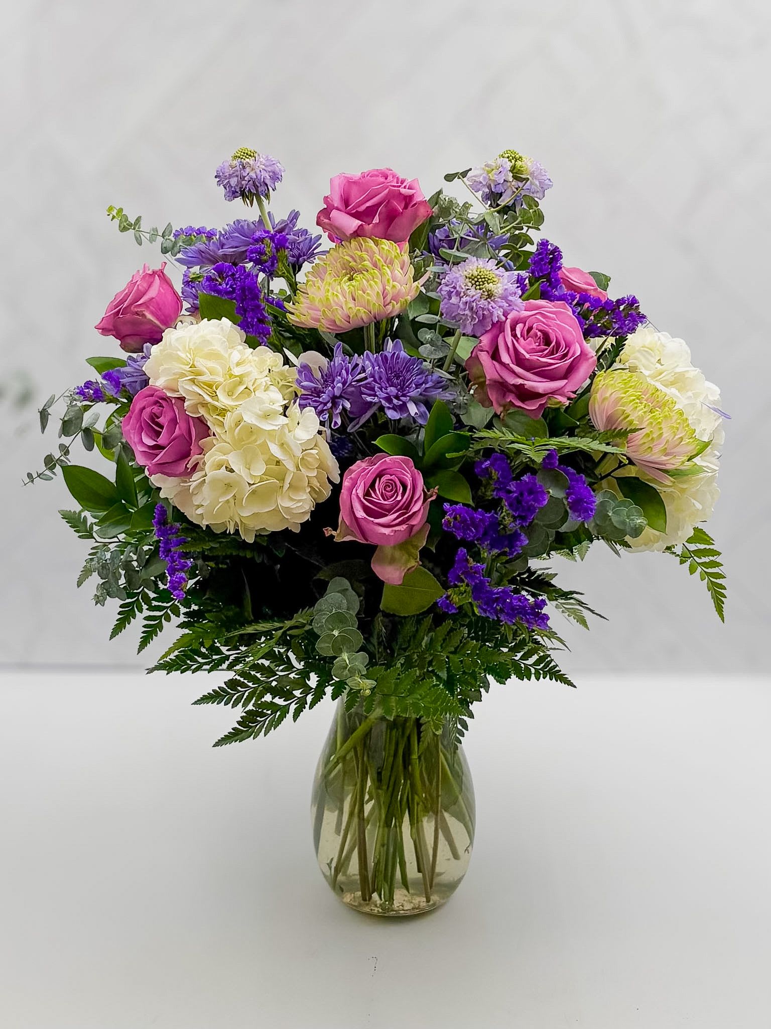 MOMS GARDEN - A lush crown of pink roses and creamy white hydrangeas makes a gentle, romantic statement-perfect as an anniversary bouquet or a birthday surprise that reads as both joyful and thoughtful. This flower arrangement, presented in a clear glass vase, is also the kind of "thinking of you" gift that comforts: the soft lavender and deep purple accents create a calming, modern contrast that will make the recipient feel truly seen.  The design combines full-headed hydrangeas, garden-style pink roses, pale green-tipped spider chrysanthemums, small lavender asters, and other seasonal fillers for a layered, textural look. The upright, rounded silhouette sits above a neat collar of greenery that frames the blooms; proportions are balanced so the bouquet looks abundant without feeling fussy. The clear glass vase lets stems show through for a fresh, contemporary presentation.  Send this arrangement to mark a milestone or brighten an ordinary day-she'll feel celebrated whether it's a birthday, anniversary, or a simple "I'm thinking of you." For best enjoyment, place in fresh water away from direct heat, and refresh the water every two days to help the blooms stay vibrant. Add a little something extra, gourmet chocolates, hand poured candles. See all our gift selections under the thoughtful gifts tab. Or call the shop and let us know what you are looking for and we will do the shopping for you.