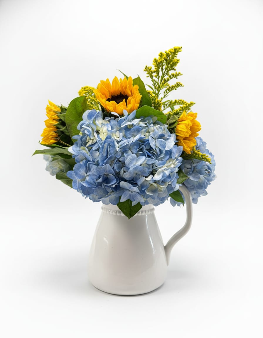 SUNNY SKIES - Blue hydrangeas form a lush, rounded mound punctuated by tall, sunny sunflowers-an uplifting birthday bouquet or thank-you arrangement that reads cheerful from across the room. The cool, pale blues and crisp whites balance the warm yellow centers and green foliage, so the person you're thinking of will feel brightened and genuinely celebrated the moment they see it.  Arranged in a white ceramic pitcher-style vase, the composition feels simple and homey while still looking abundant and thoughtfully composed. Small yellow filler blooms add airy height above the hydrangea mass, giving the piece light texture and visual movement. Perfect for everyday gifting, summer decorating, or casual celebrations; the compact, mound-and-spray design makes it easy to place on a table, counter, or mantel-no extra styling required.