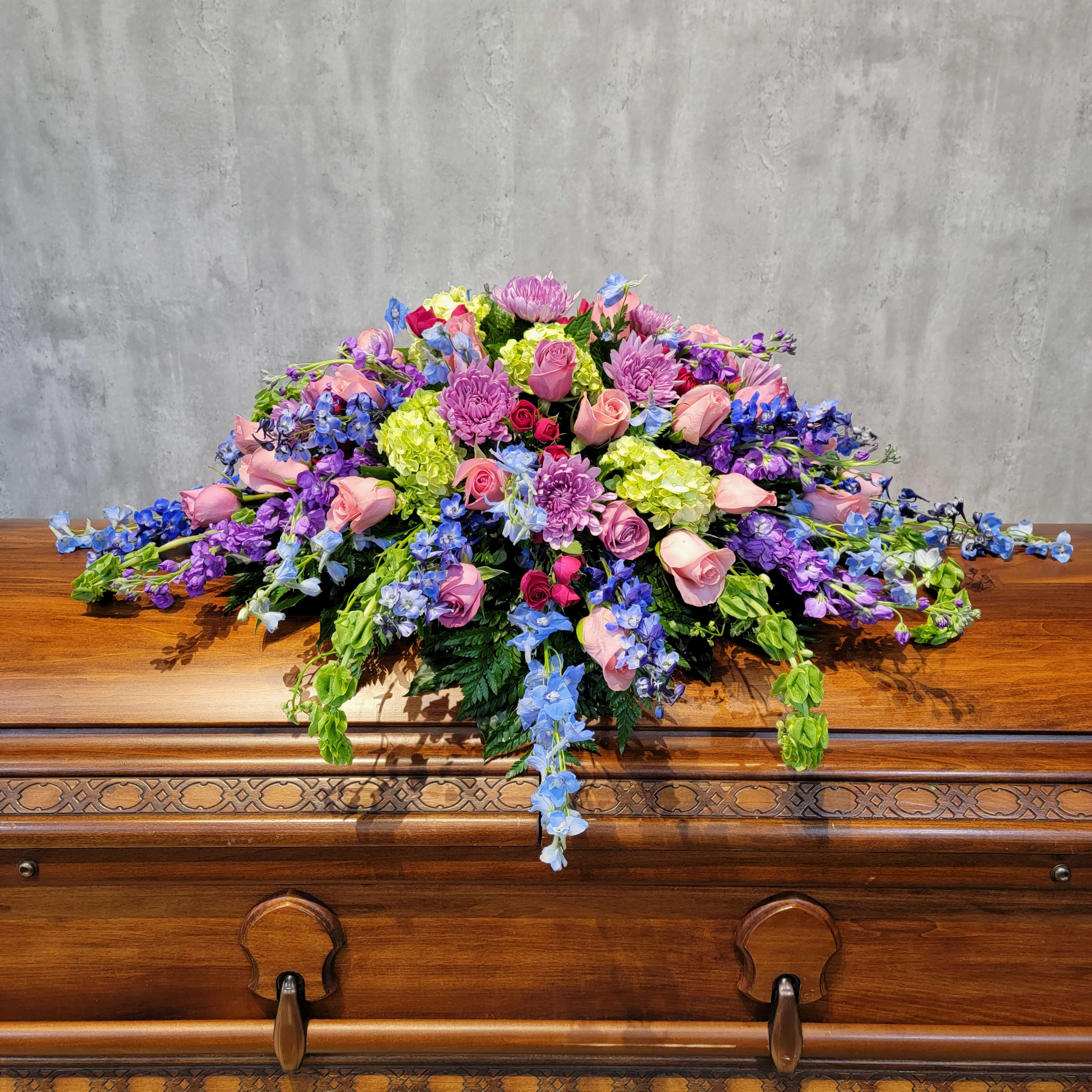 Large floral spray with pink roses, purple blooms, and blue flowers on a wooden casket