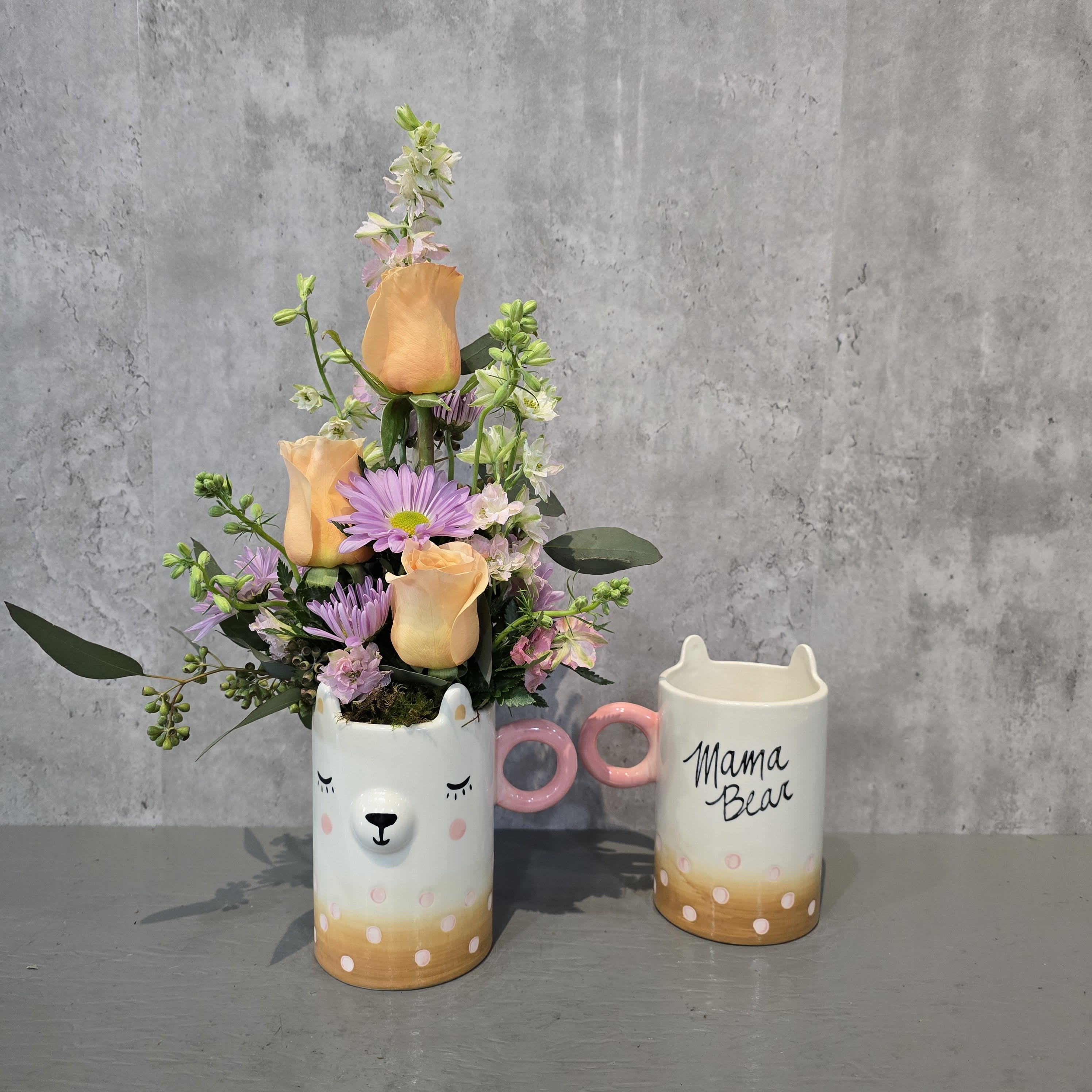 Peachy Mama - Sweet, soft, and full of charm, Peachy Mama is a floral arrangement designed to warm hearts and brighten days. Featuring peach roses, lavender daisies, soft larkspur, and touches of eucalyptus, this handcrafted bouquet blooms from a whimsical ceramic mug with a sleepy bear face on one side and “Mama Bear” on the back. Perfect for Mother’s Day, celebrating new moms, or sending cozy love to someone special, this unique gift is available for flower delivery throughout Albuquerque, New Mexico.