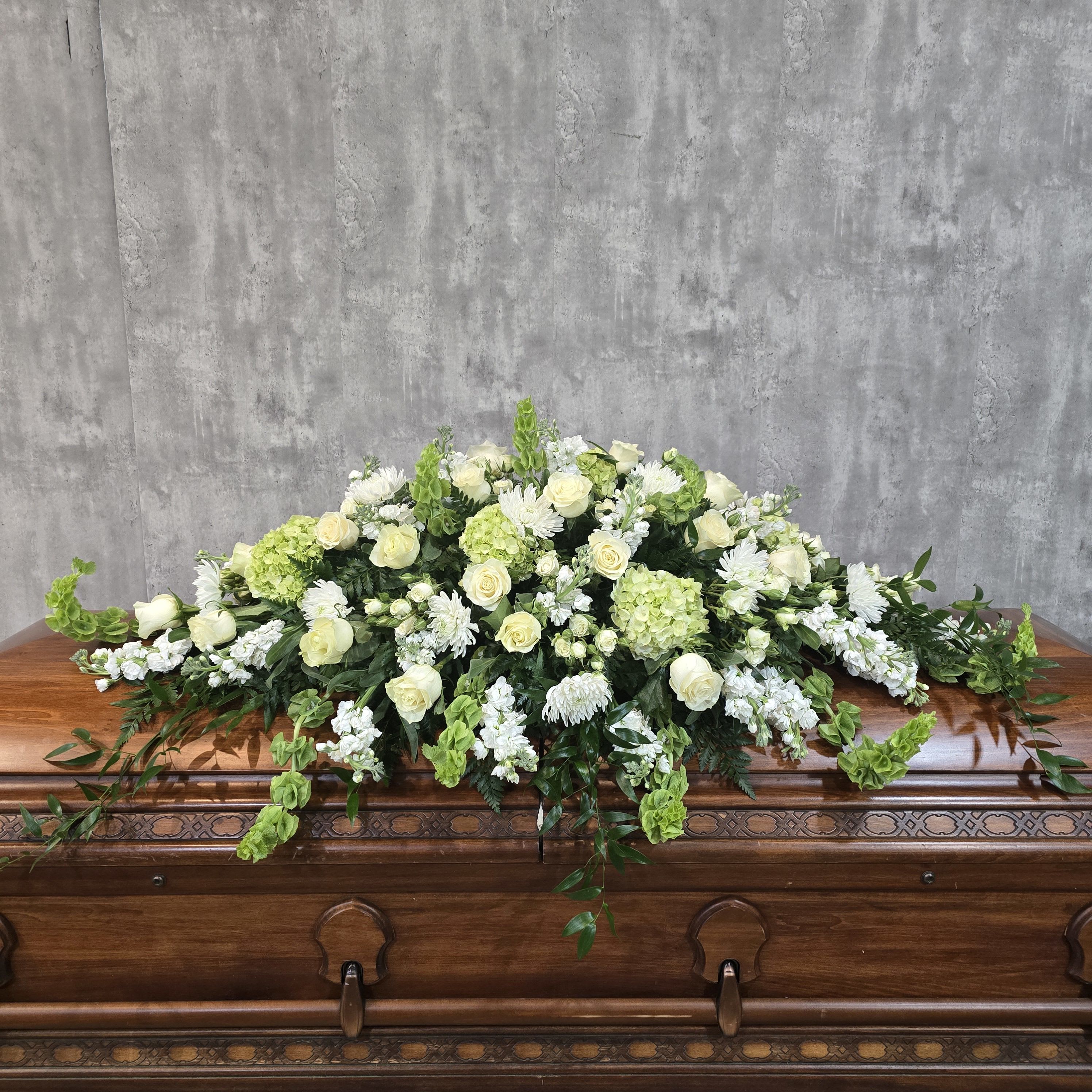 Pure Elegance Casket Spray - A low, broad casket spray crafted to convey calm and reverence-this funeral flower arrangement pairs white roses and clusters of white stock with pale green hydrangeas and white chrysanthemums for a pure, understated tribute. As a casket spray for a memorial or funeral service, it reads as traditional and elegant, offering a comforting visual that helps the person you're honoring feel remembered with grace.  The composition is large and gently cascading, arranged to flow over the casket with trailing greenery that extends beyond the main body of the spray for a soft, layered silhouette. Textures move from the dense blooms of hydrangea and chrysanthemum to the refined petals of roses and the delicate spikes of stock, all in a white-and-green palette that emphasizes serenity and respect.  This piece is intended to rest atop the casket and presents a formal, sympathetic statement appropriate for close family or as an expression of deep condolence. Its generous size and classic color scheme make it suitable for traditional services-please let us know service timing and delivery details so we can ensure fresh placement on the day.