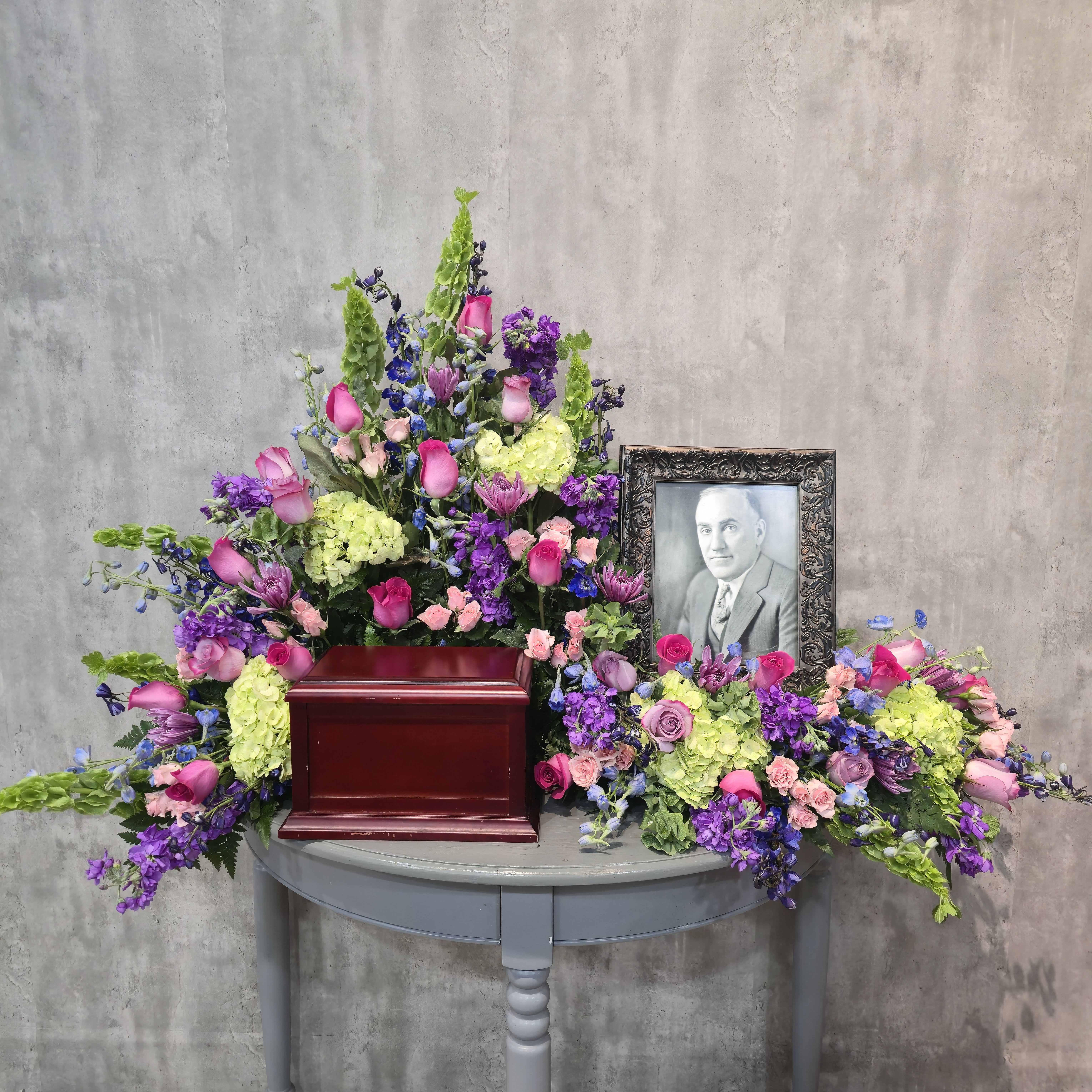 Spring Rain Urn and Photo spray - A lush sympathy flower arrangement built for an urn and photo (8x10 or 11x14), perfect as a memorial spray to honor a loved one. This funeral urn and photo spray combines pink roses, pale green hydrangeas, blue delphinium and purple stock, and touches of light pink and deeper jewel-toned blooms so the person you're remembering will feel celebrated with dignity and warmth. The framed portrait beside the urn and the broad, cascading shape give the piece a formal, commemorative presence suited to a visitation or funeral service.  The composition is generous and layered: soft pink roses provide a tender focal point, tall blue delphinium adds vertical movement, and pale green hydrangeas fill the design for a lush, textural base. Accents of stock, and other seasonal blooms create color depth between the pastels and richer hues brings a natural, understated gravity to the display. Sized to drape gracefully over a table, the spray reads as abundant without being overpowering.  Designed to comfort and to honor, this arrangement is an appropriate choice for family services, memorials, or when you want to include a framed portrait alongside flowers. Ask us about delivery to the funeral home or chapel and any personalization-we'll work with you to ensure the spray's scale and color balance match the service and the person you're commemorating.