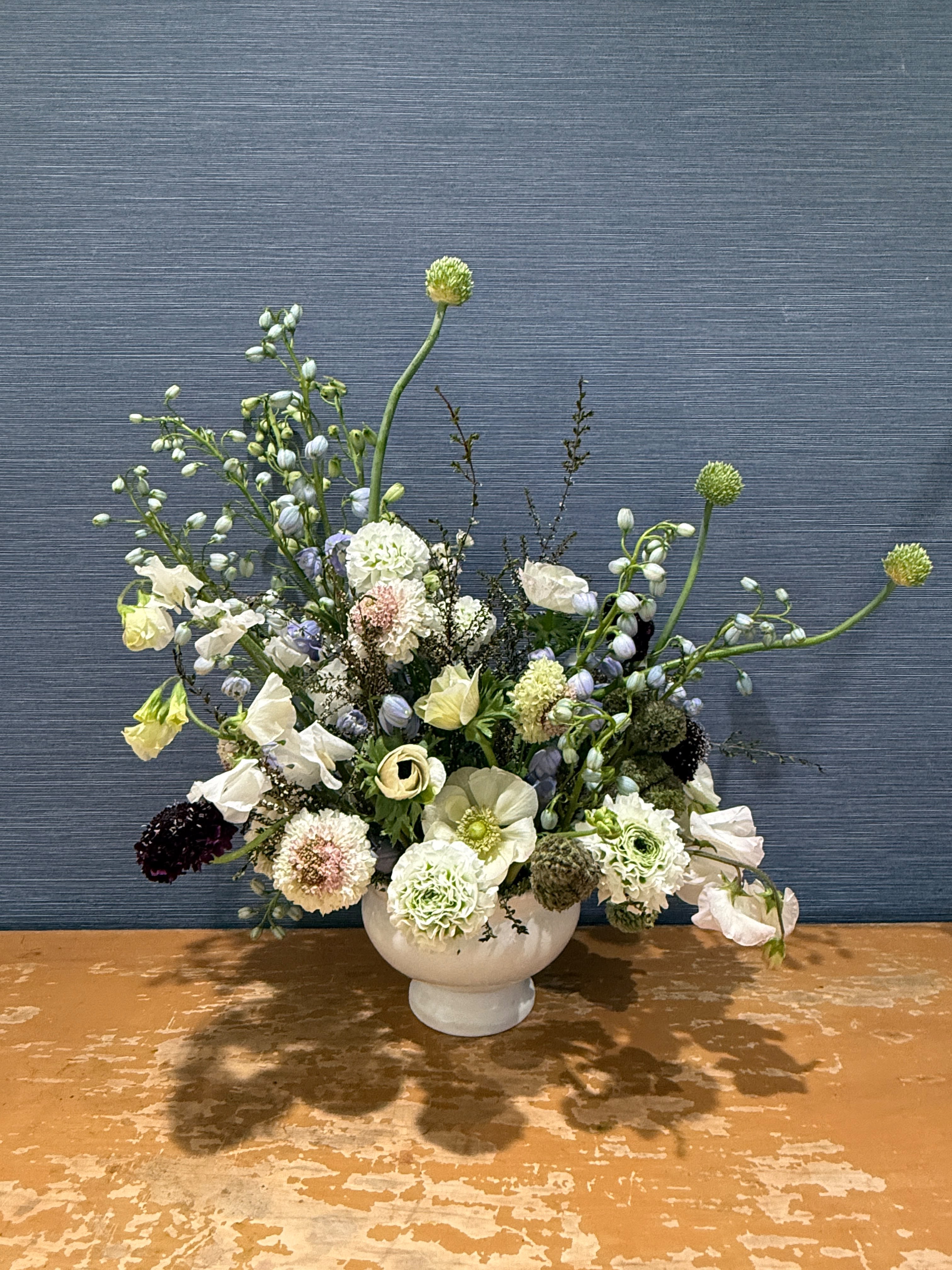Meadow &amp; Mist  - A lush, garden-gathered arrangement in a white ceramic compote featuring white anemones, cream ranunculus, green-edged flowers, blush and burgundy scabiosa, and soft lavender delphinium buds — with wild reaching stems and wispy branches adding movement