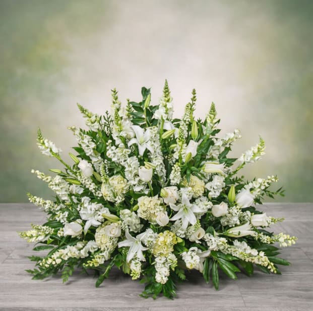 Elegant White Floor Basket - A serene fan-shaped sympathy flower arrangement, this Elegant White Floor Basket communicates calm and reverence for memorial services or quiet ceremonial moments. The design reads formal and dignified-an ideal sympathy arrangement or back-of-room piece for a funeral, graveside service, or a solemn wedding or religious event where understated elegance is called for.  Creamy white lilies, roses, hydrangeas, snapdragons and carnations form layered textures from a low, elongated base up to taller snapdragon spikes, while pale yellow highlights warm the cool white palette. Dense green foliage grounds the piece and gives the basket a structured, fan-like silhouette that looks graceful placed at the front of a room or beside a casket.  Give someone the comfort of a composed, respectful tribute that lets them feel honored and remembered. Designed as a floor basket, this arrangement is sized to stand on its own at services; please contact us for delivery timing and placement guidance to ensure it arrives fresh and properly positioned for the occasion.