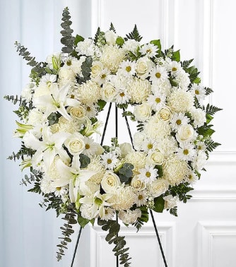 White Funeral Wreath - A large circular standing wreath of white roses, lilies, chrysanthemums and small daisy-style blooms creates a serene, comforting tribute for a funeral or memorial service. The white funeral wreath brings a calm, respectful presence to a chapel, graveside, or service-an elegant way to express sympathy and honor the person you're thinking of.  Flowers are densely clustered into a full ring with subtle ivory and pure-white tones; darker green foliage and a few trailing stems add contrast and a gentle cascading effect at the bottom. The arrangement is mounted on a sturdy metal wreath frame and shown on a tall black metal easel for formal display, giving it a polished, dignified presentation.  This wreath is meant to convey sympathy and reverence-an appropriate choice for close family, dear friends, or formal services where a classic, timeless tribute is called for. Carefully wired to hold shape during transport and display; please let us know the service location and timing so we can deliver and set the easel in place.