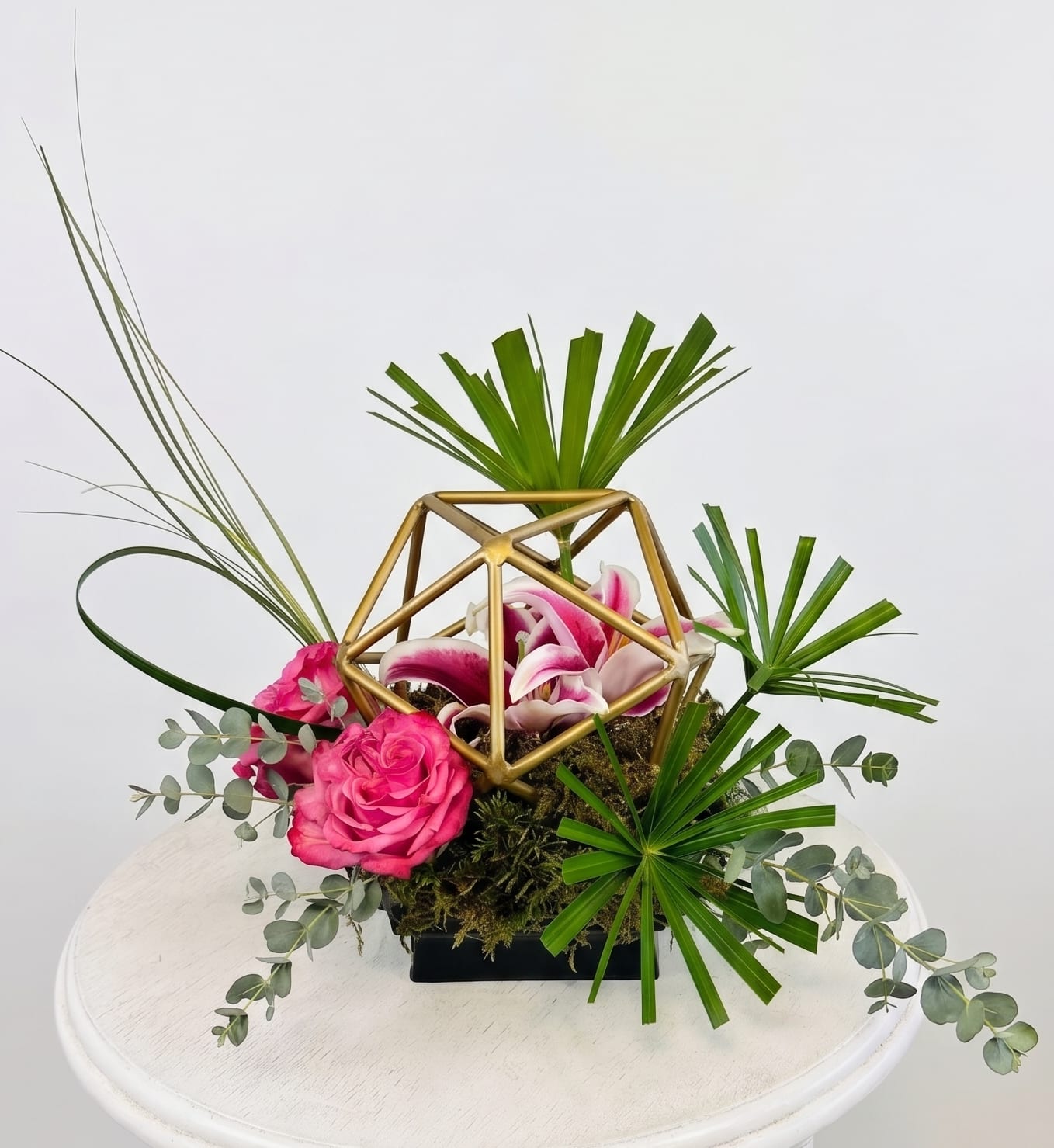 Golden Prism Bouquet - Artistry and architecture collide in the breathtaking Golden Prism Bouquet. Centered around a gleaming, open-faceted gold geometric prism, this avant-garde arrangement elevates traditional floristry into a modern work of art.  Nestled within the metallic structure is a striking, pink-trimmed lily resting on a bed of rich forest moss. Radiating outward are brilliant magenta-pink roses, structured fan palms, and delicate, cascading sprigs of silver-dollar eucalyptus that offer a beautiful, organic contrast to the sharp lines of the prism.  Finished in a sleek, low-profile black container and styled to perfection on a clean white surface, this sculptural masterpiece is the ultimate statement piece.   It is perfect for contemporary home decor, upscale corporate events, or as an unforgettable gift for someone who appreciates bold, sophisticated design.