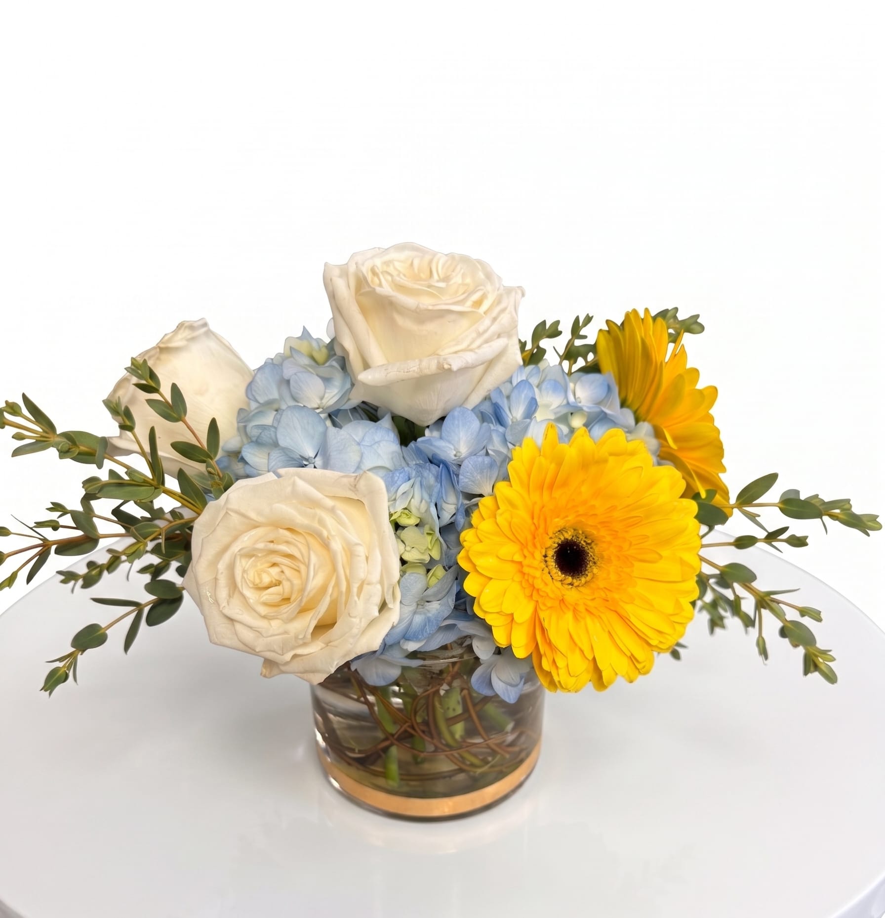 Sunny Skies Bouquet - Bring a splash of pure joy and warmth into any room with this stunning arrangement.  Beautifully balanced and bursting with cheer, this design features vibrant yellow gerbera daisies nestled against a soft, cloud-like bed of serene blue hydrangeas. Elegant white roses add a touch of classic sophistication, while wisps of fresh eucalyptus give it a natural, airy movement.  Arranged in a sleek, modern glass cylinder vase, this piece is perfectly sized for a coffee table, office desk, or a cozy dining setup. It’s the ultimate way to send a little sunshine, celebrate a special day, or simply say, "I'm thinking of you."