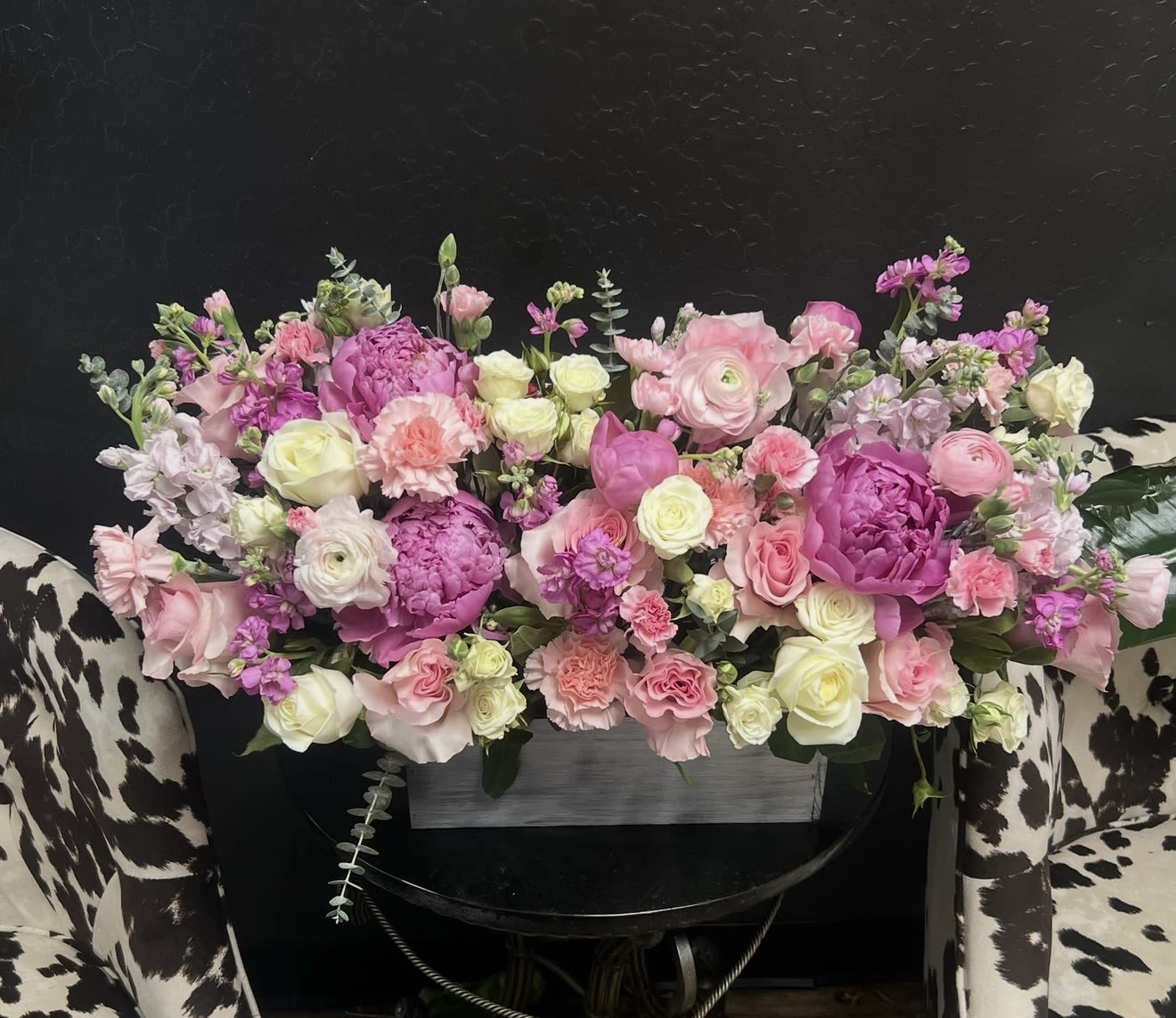 Pink Peony and Ranunculus Love Centerpiece  - Lush pink peonies and soft ranunculus create a romantic, garden-fresh centerpiece perfect for a wedding tablescape or anniversary celebration. This pink peony and ranunculus love centerpiece places the flowers low and wide-a large, low centerpiece or wedding centerpiece-so the person you're celebrating can still talk across the table while feeling surrounded by blush, cream, lavender, and green petals.  The design mixes rounded focal peonies and roses with clusters of ranunculus, spray roses, carnations, and stock for a layered, textured look. Presented in a rectangular wooden container, the arrangement spreads broadly with a few airy stems rising above the main mass and trailing eucalyptus-like foliage that softens the edges. Colors include pink, white, purple, and green for a pastel palette that reads soft and romantic.  For weddings, anniversaries, or spring gifting, this piece reads as celebratory and intimate-the kind of centerpiece that makes the recipient feel truly seen and cherished. Sized to cover the length of a standard banquet centerpiece area, it arrives ready to display; keep blooms in fresh water and away from direct heat to enjoy the fullest, longest-lasting look.