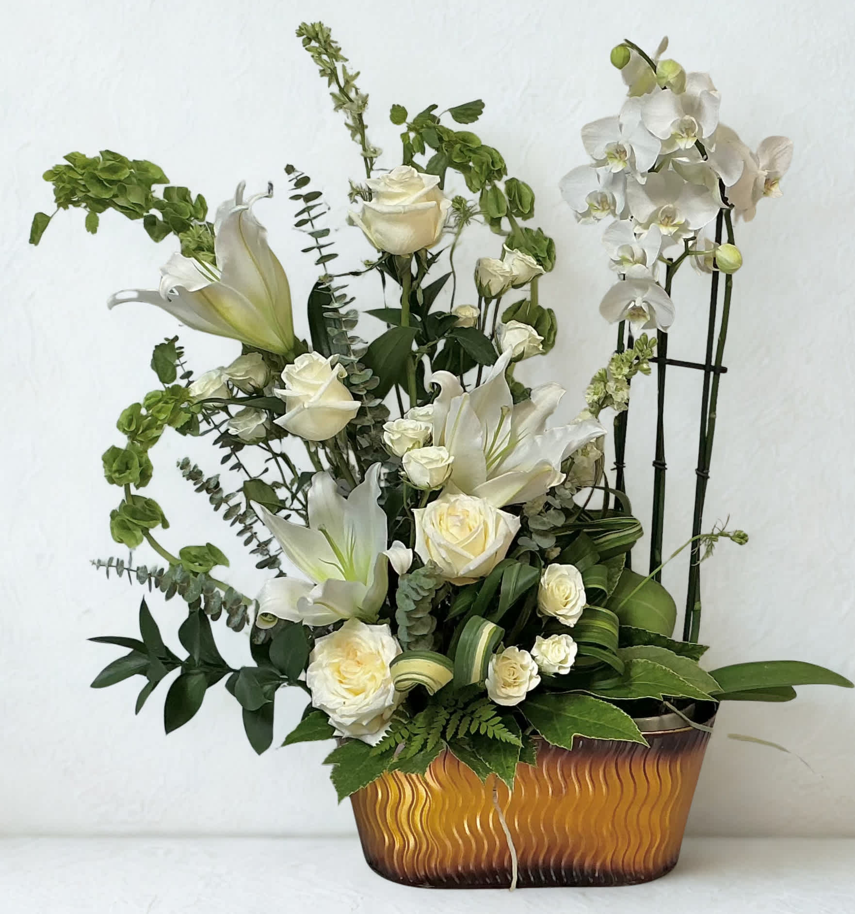 White roses and lilies arranged in a gold-toned container with orchids.