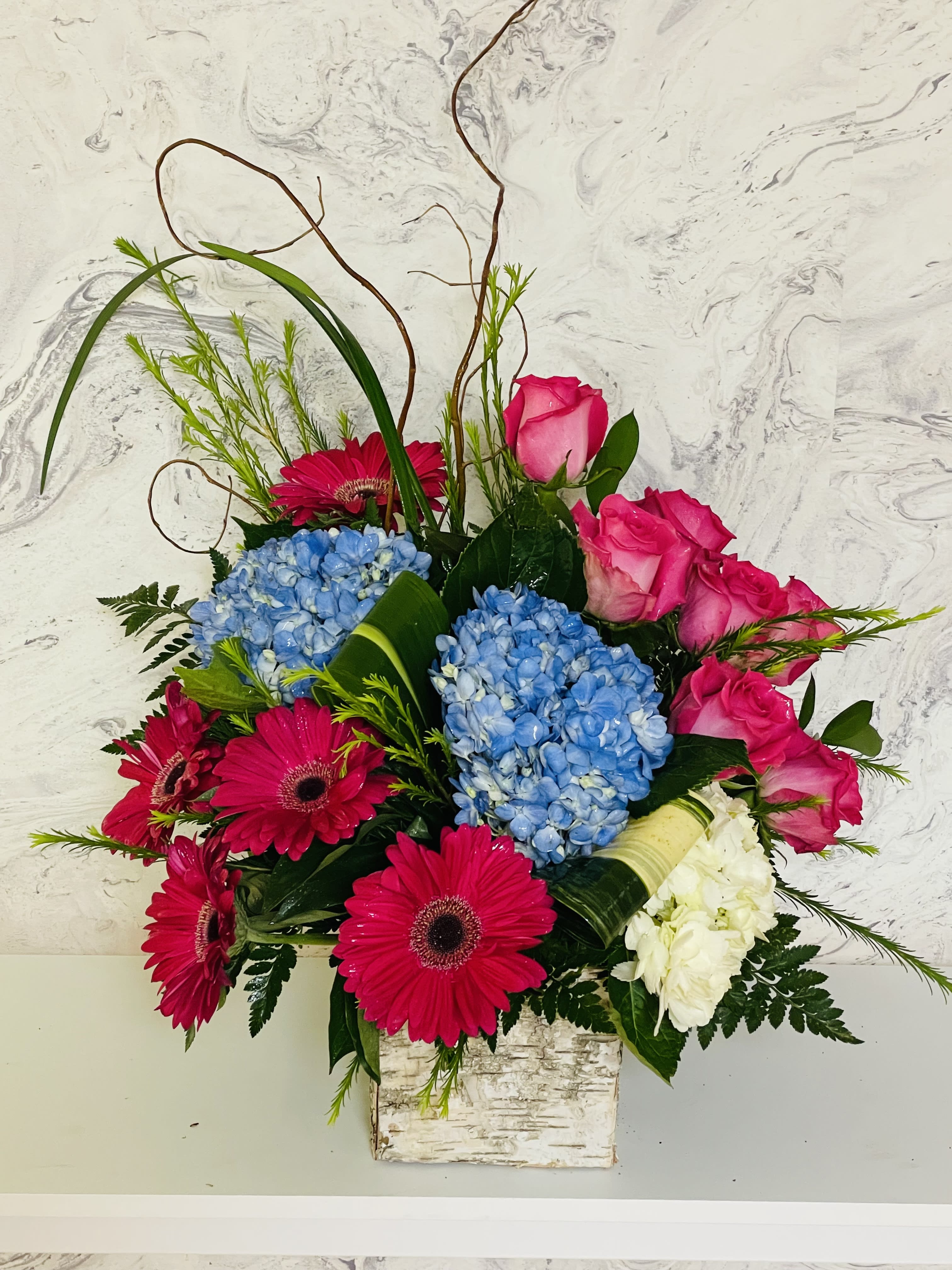 Bright bouquet of pink roses, blue hydrangeas, and magenta gerbera daisies in a square vase